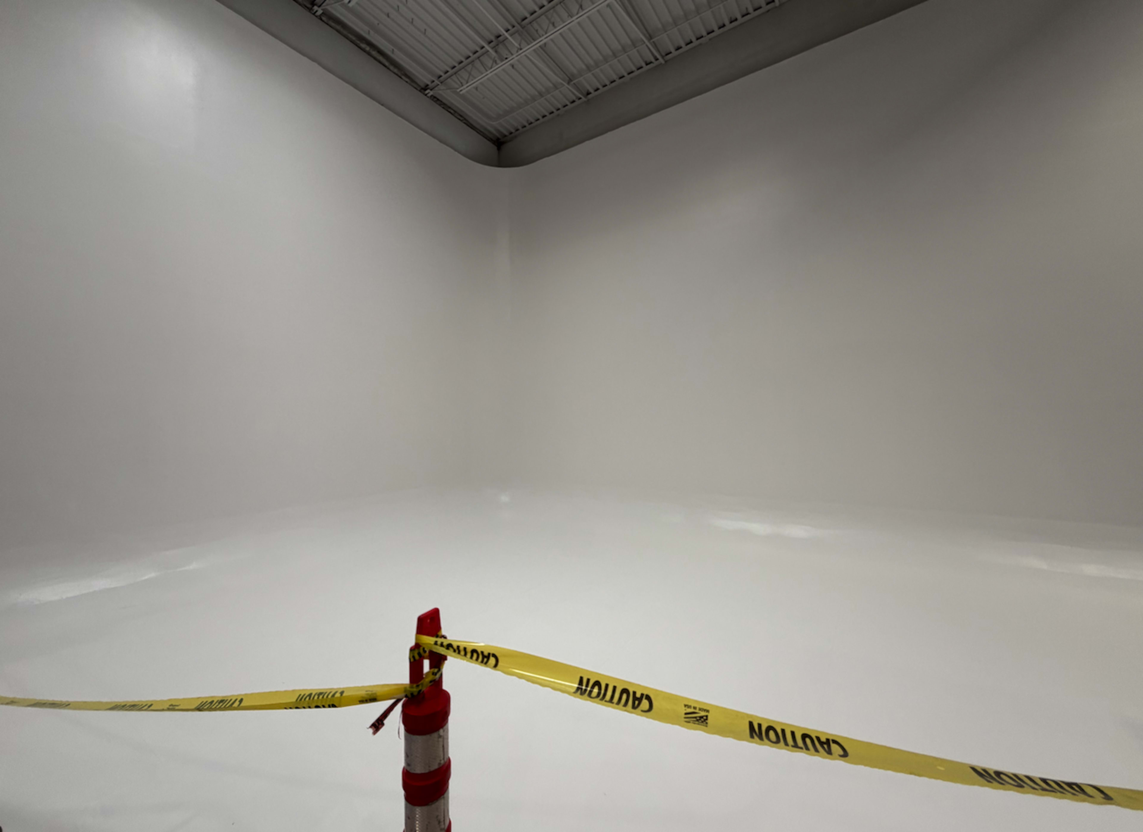 The image shows an empty, freshly painted white room with caution tape and a traffic cone in the foreground.