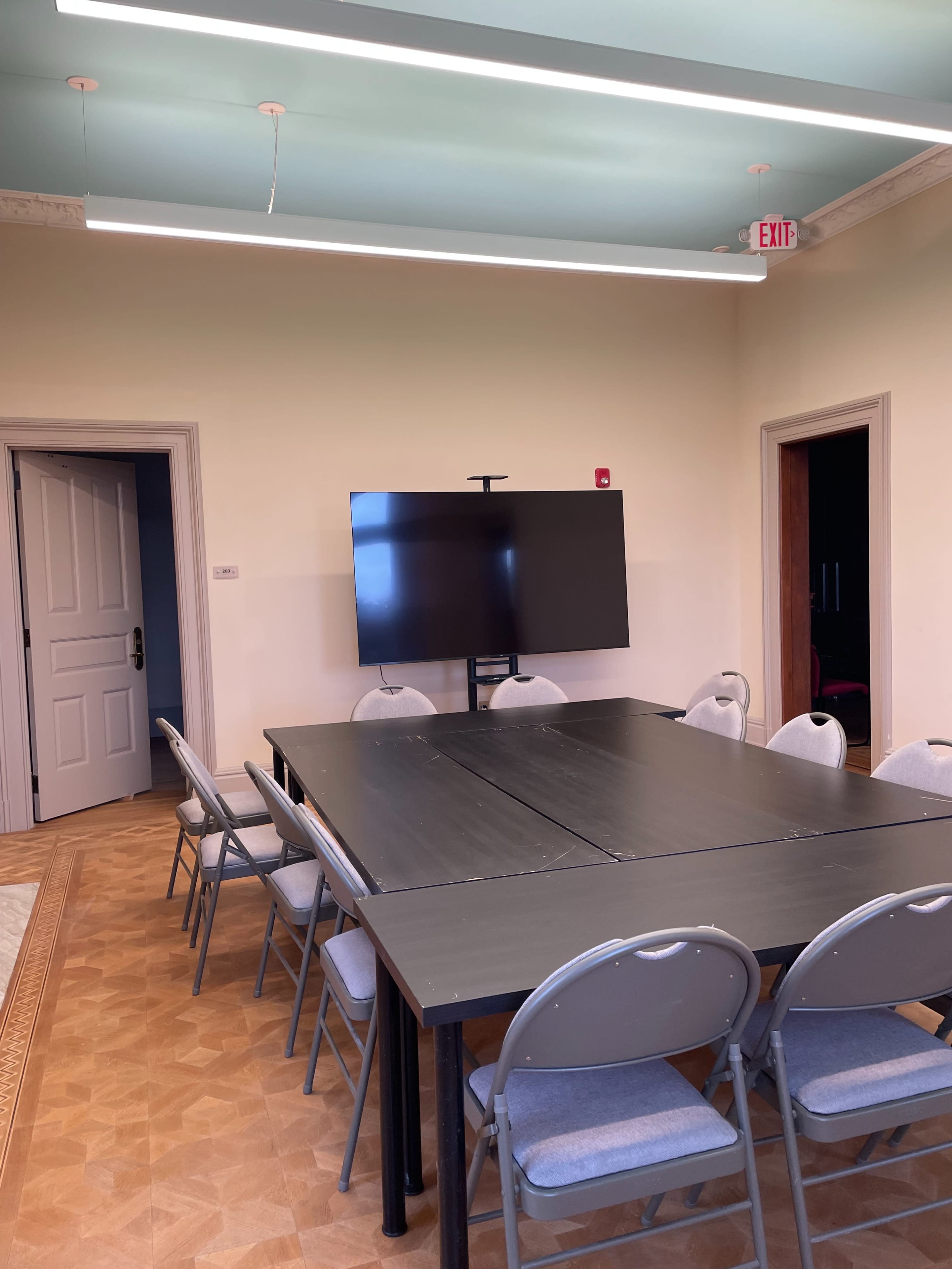 The image shows a meeting room with a large table surrounded by gray chairs and a wall-mounted flat-screen television.