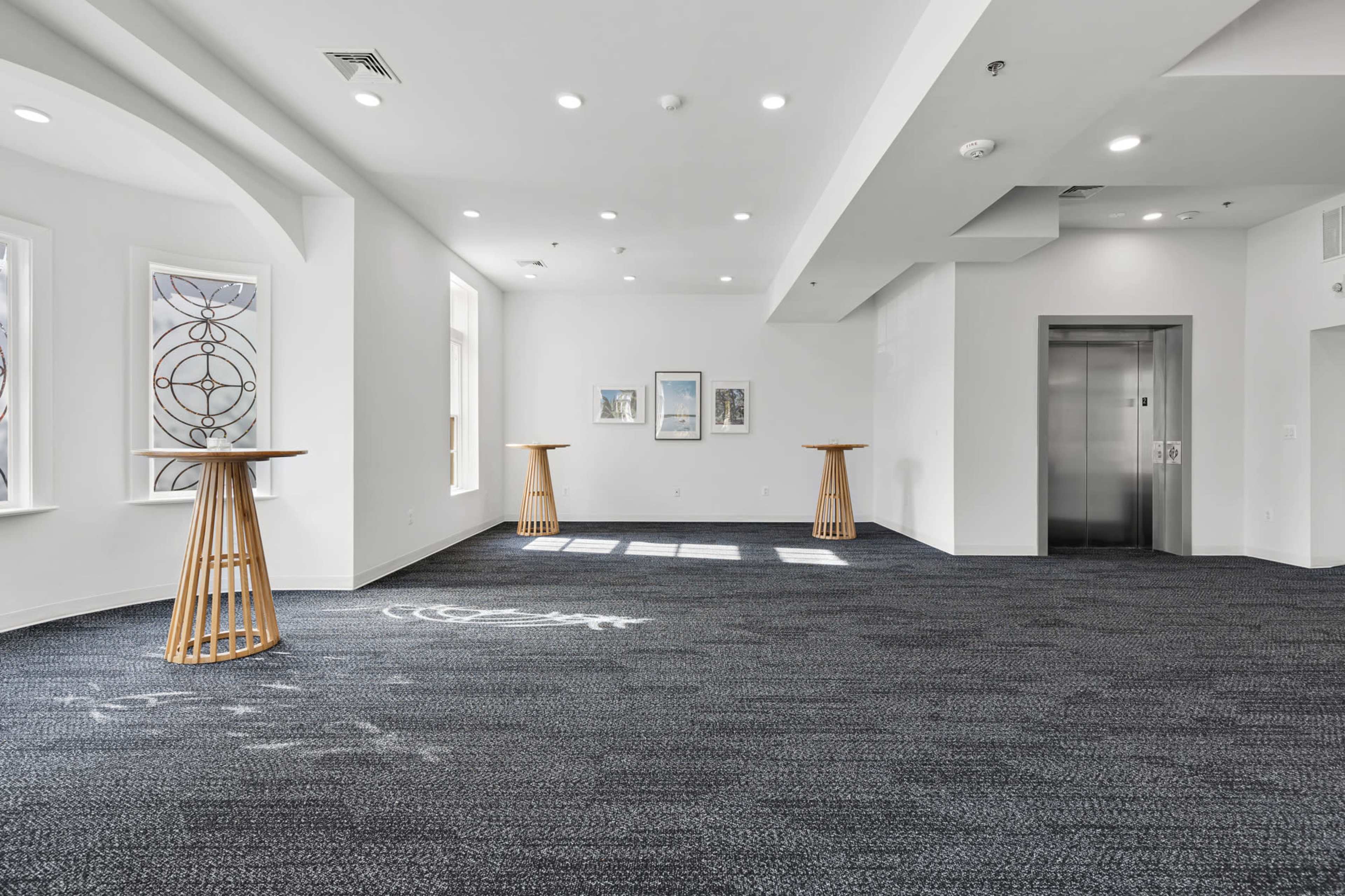 A spacious room with two wooden tables placed on a dark carpet, featuring large windows and an elevator at one end.