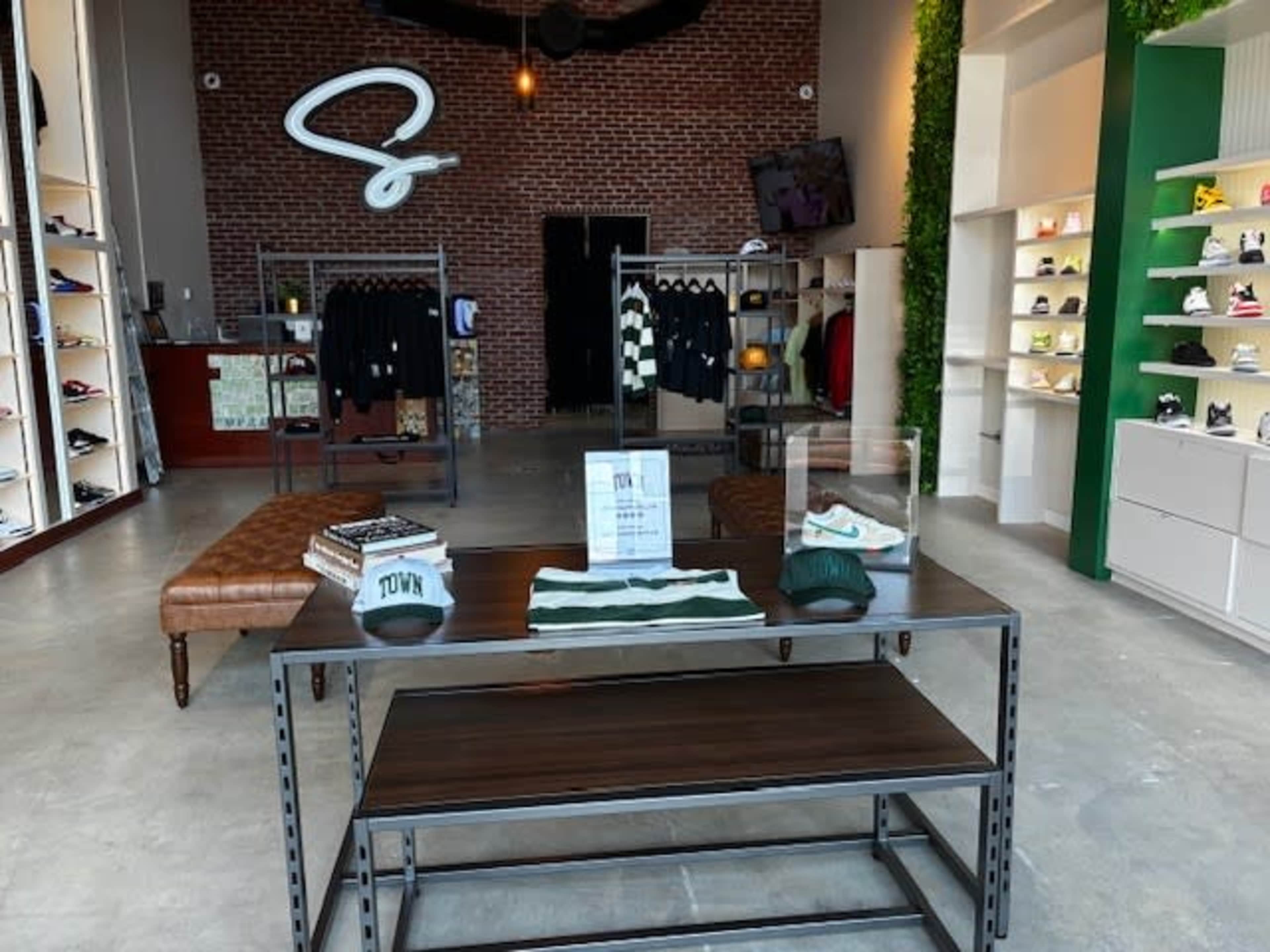 The image shows a modern retail store featuring clothing and footwear displays, with a brown leather couch and a table showcasing hats and apparel in the center.