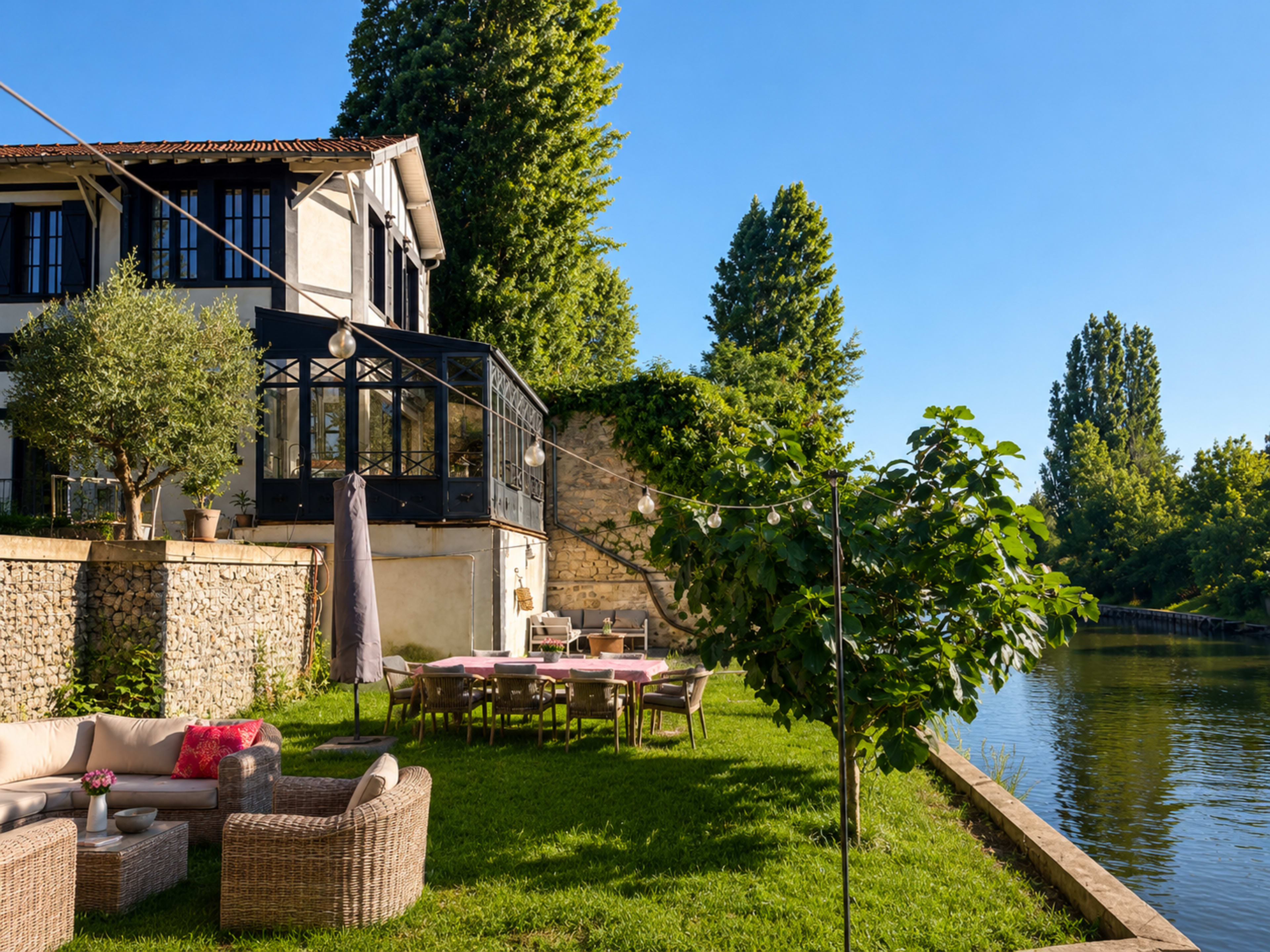 A modern house with a glass extension overlooks a calm river, surrounded by green grass and trees.