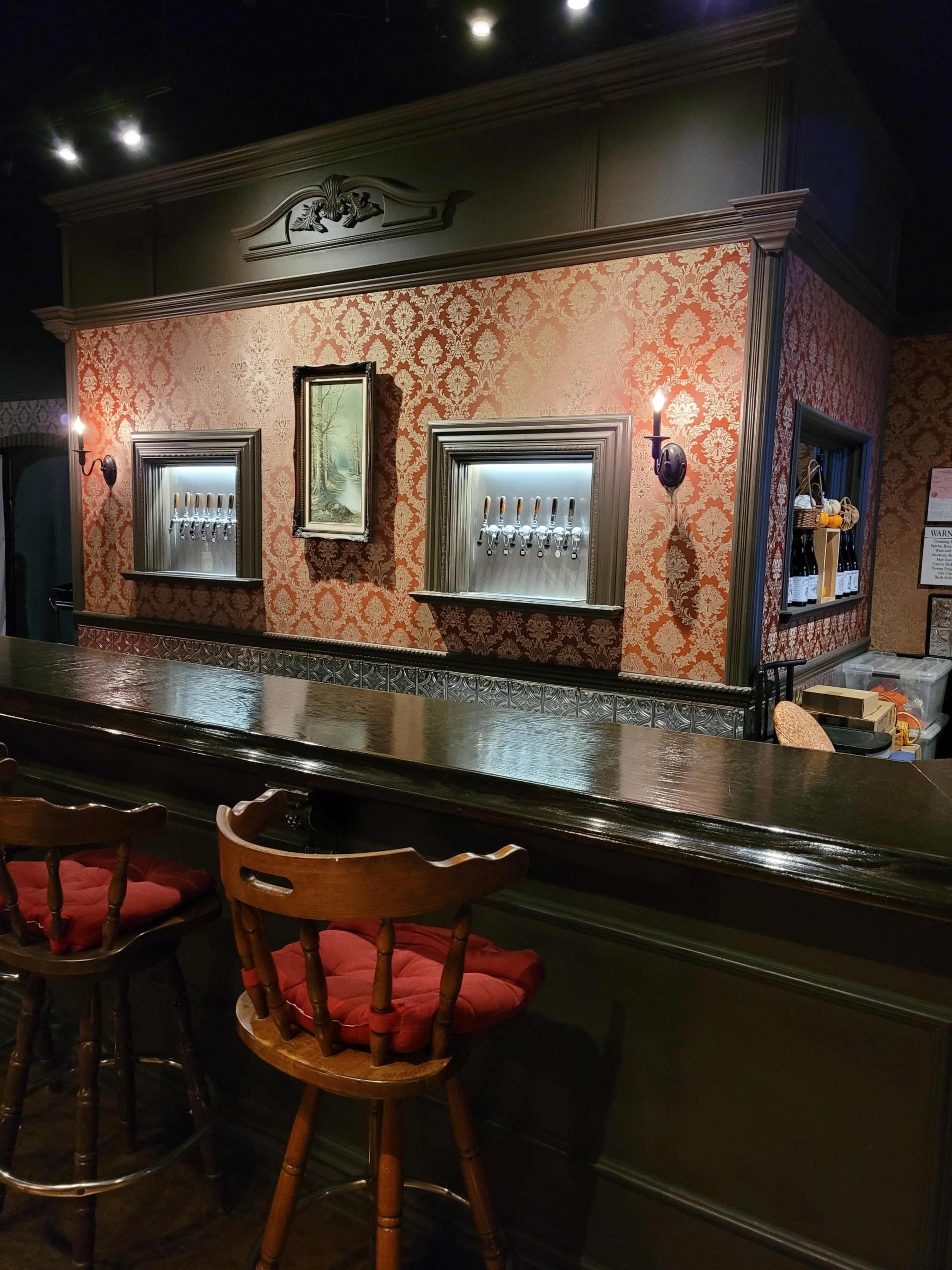 A bar area with a wooden counter, high stools, and ornate wallpaper featuring framed art and illuminated tap handles on the wall behind it.