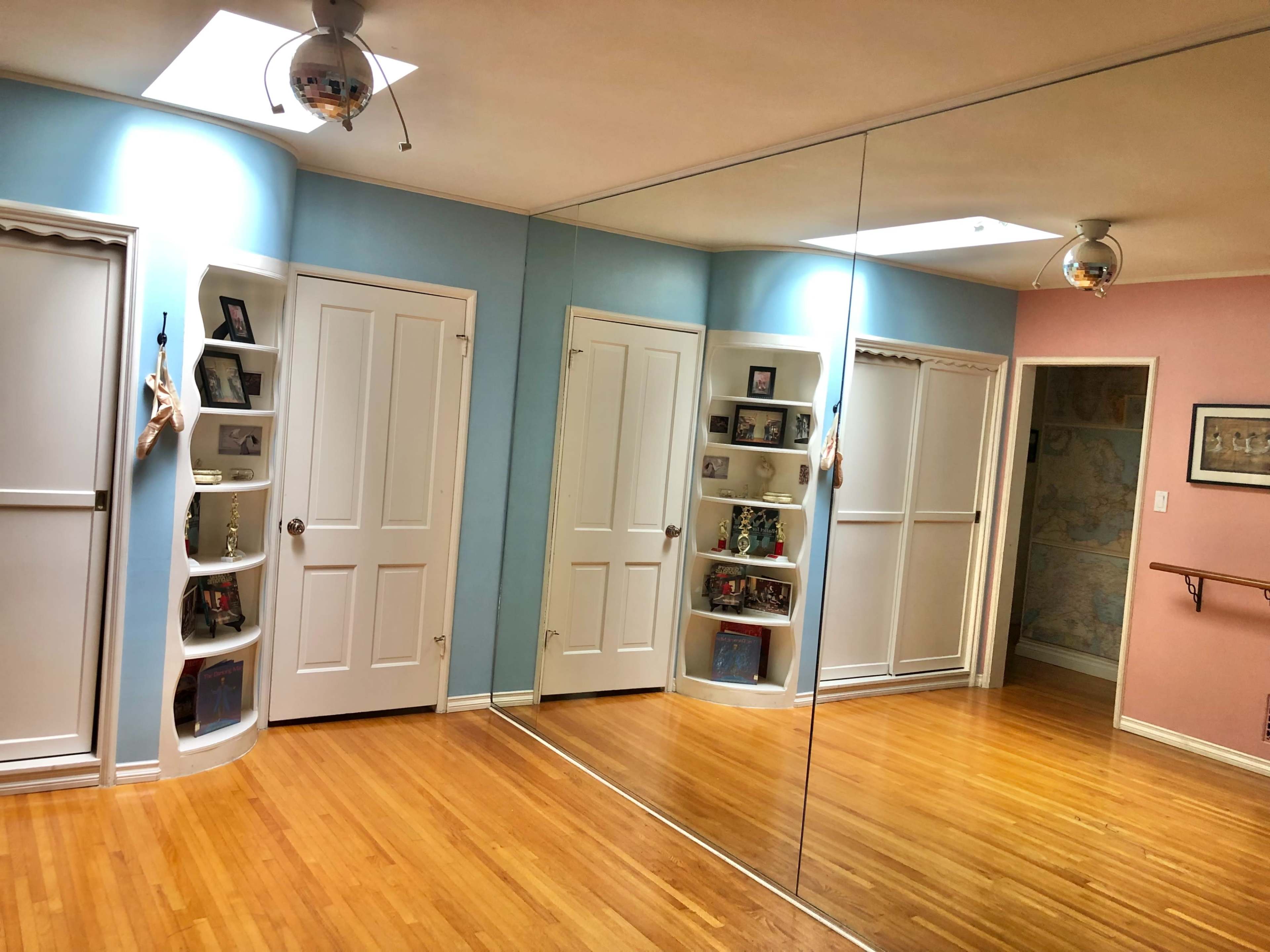 The image shows a brightly lit room with a mirrored wall, featuring wooden flooring and two doors flanked by built-in shelves on one side.