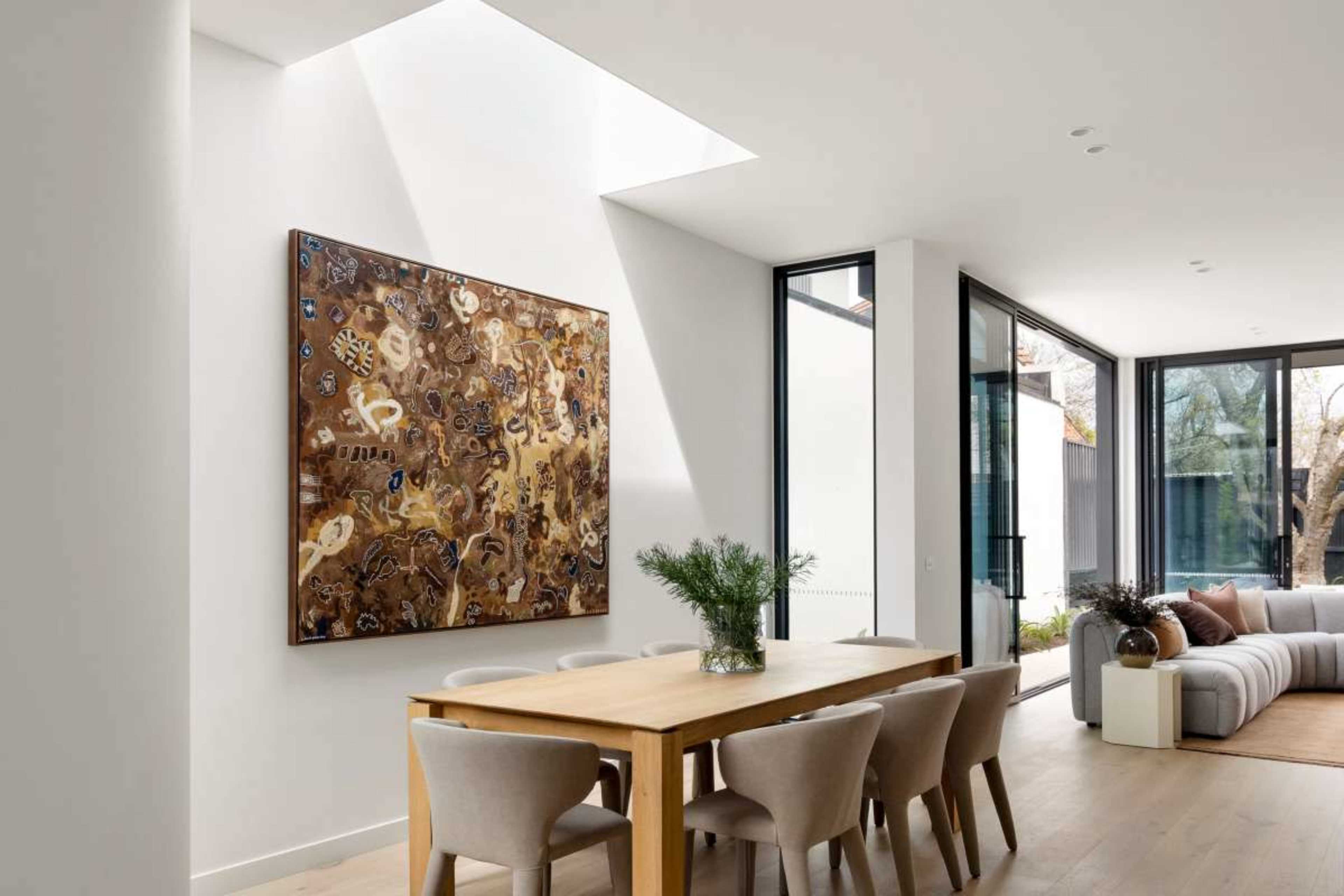 A modern dining area features a wooden table surrounded by upholstered chairs, with a large artwork mounted on the wall and natural light streaming through a skylight.