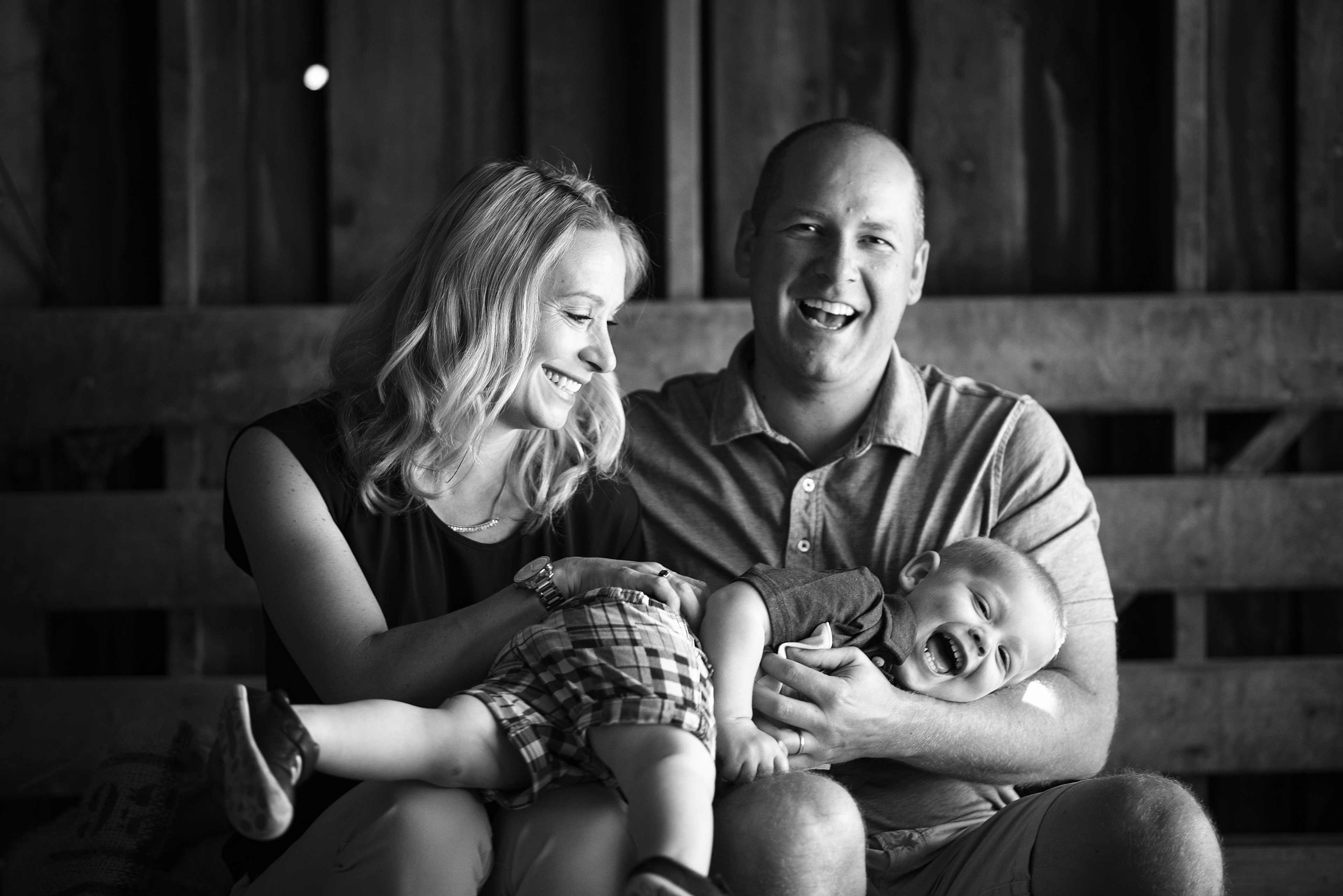 A family of three sits on a wooden bench in a barn, with the mother and father playing with their laughing toddler.