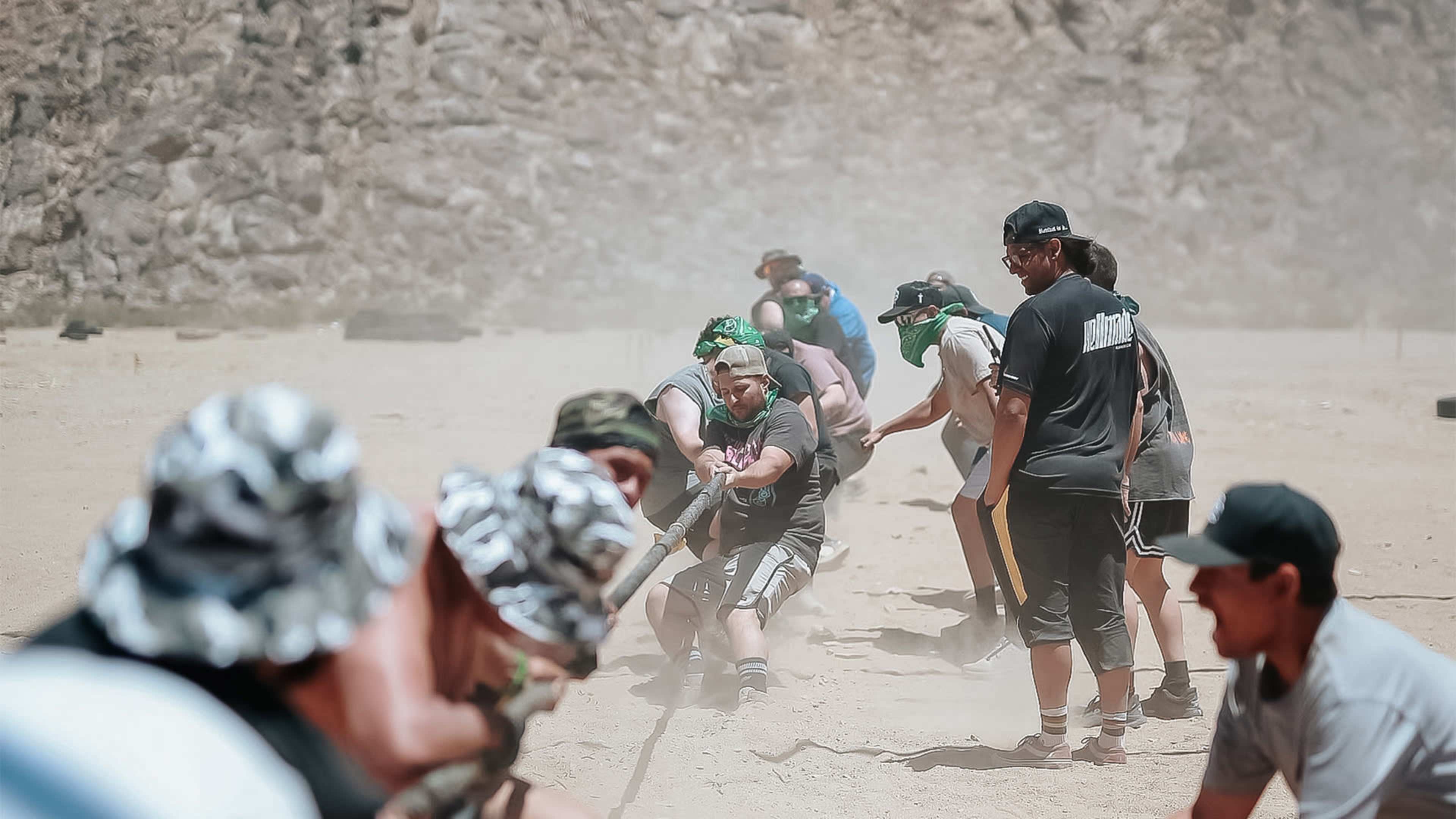 A group of people is engaged in a tug-of-war competition on a dusty terrain surrounded by rocky hills.