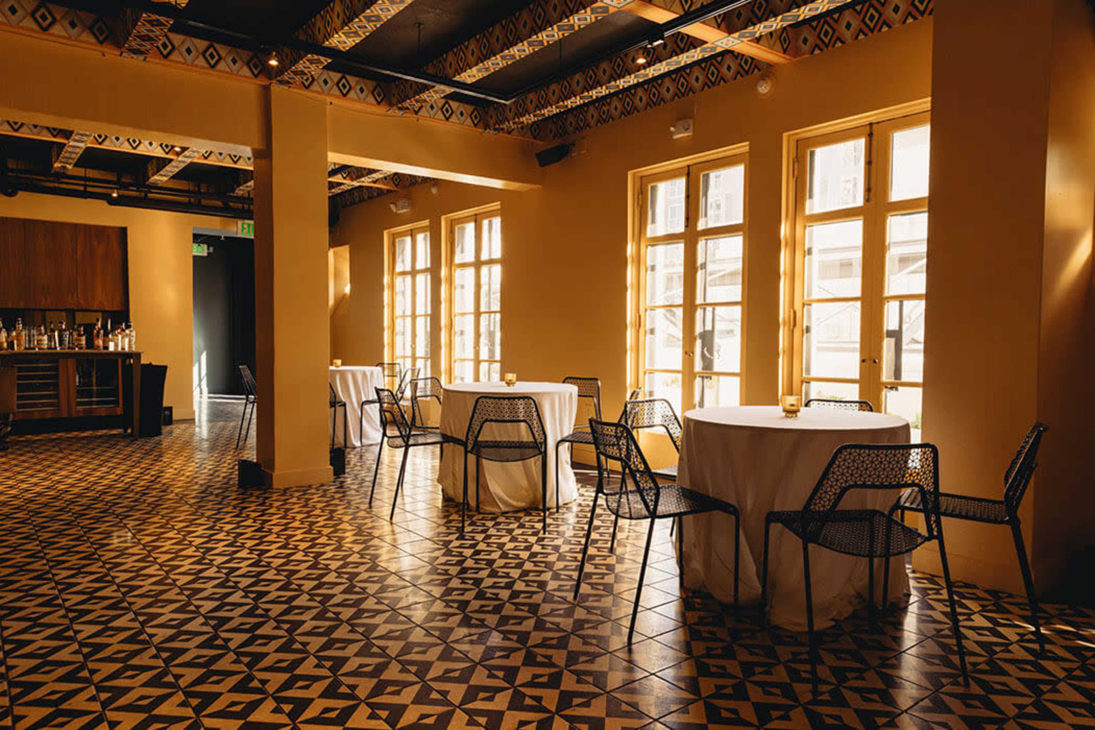 The image shows a restaurant interior with round tables covered in white tablecloths and black wire chairs arranged on a patterned tile floor.