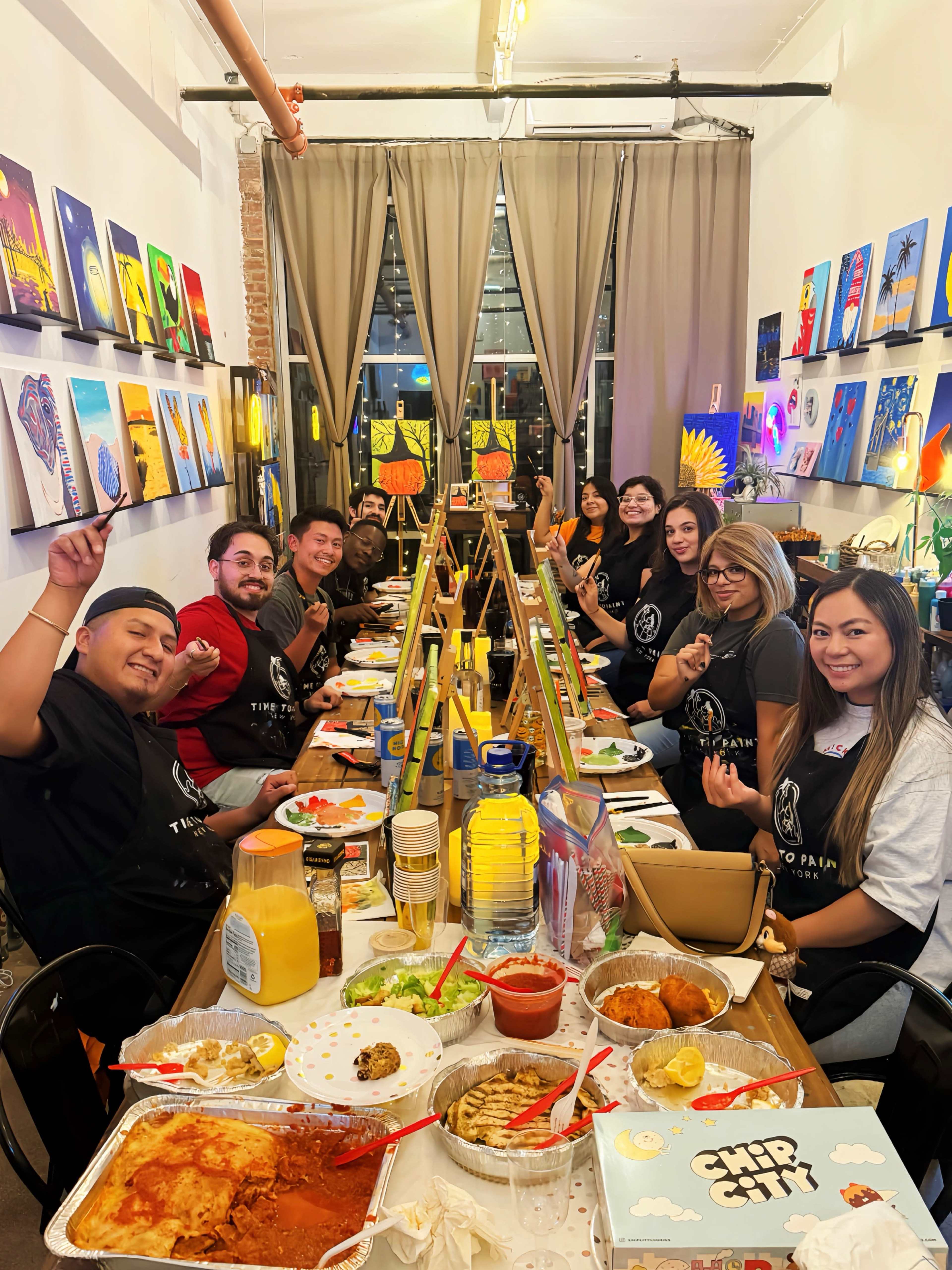 A group of twelve people is seated at a long table in a brightly lit art studio, enjoying a meal while surrounded by colorful paintings hanging on the walls.