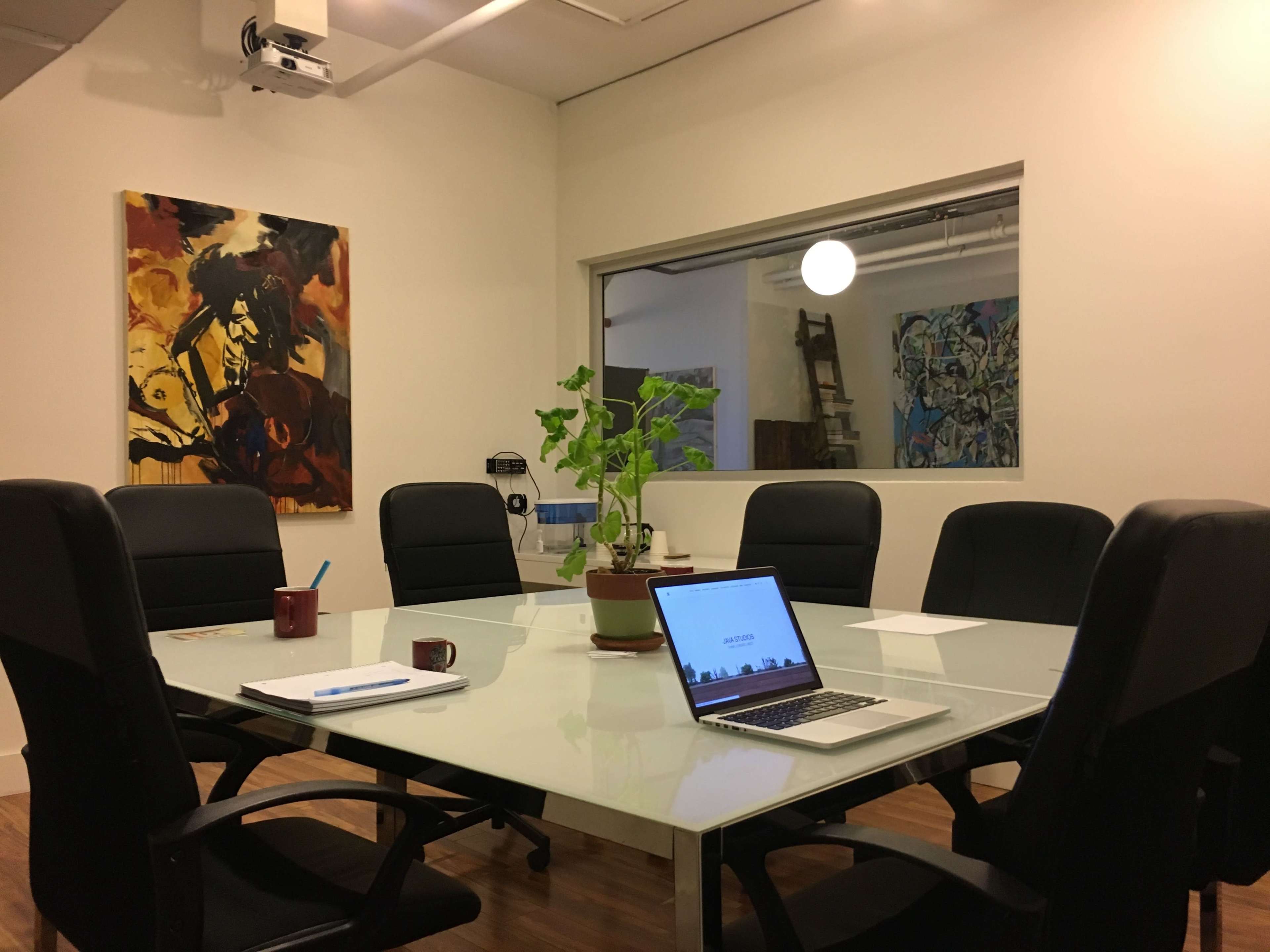 A modern conference room features a glass table surrounded by black chairs, a laptop, a plant in the center, and two artworks on the walls.