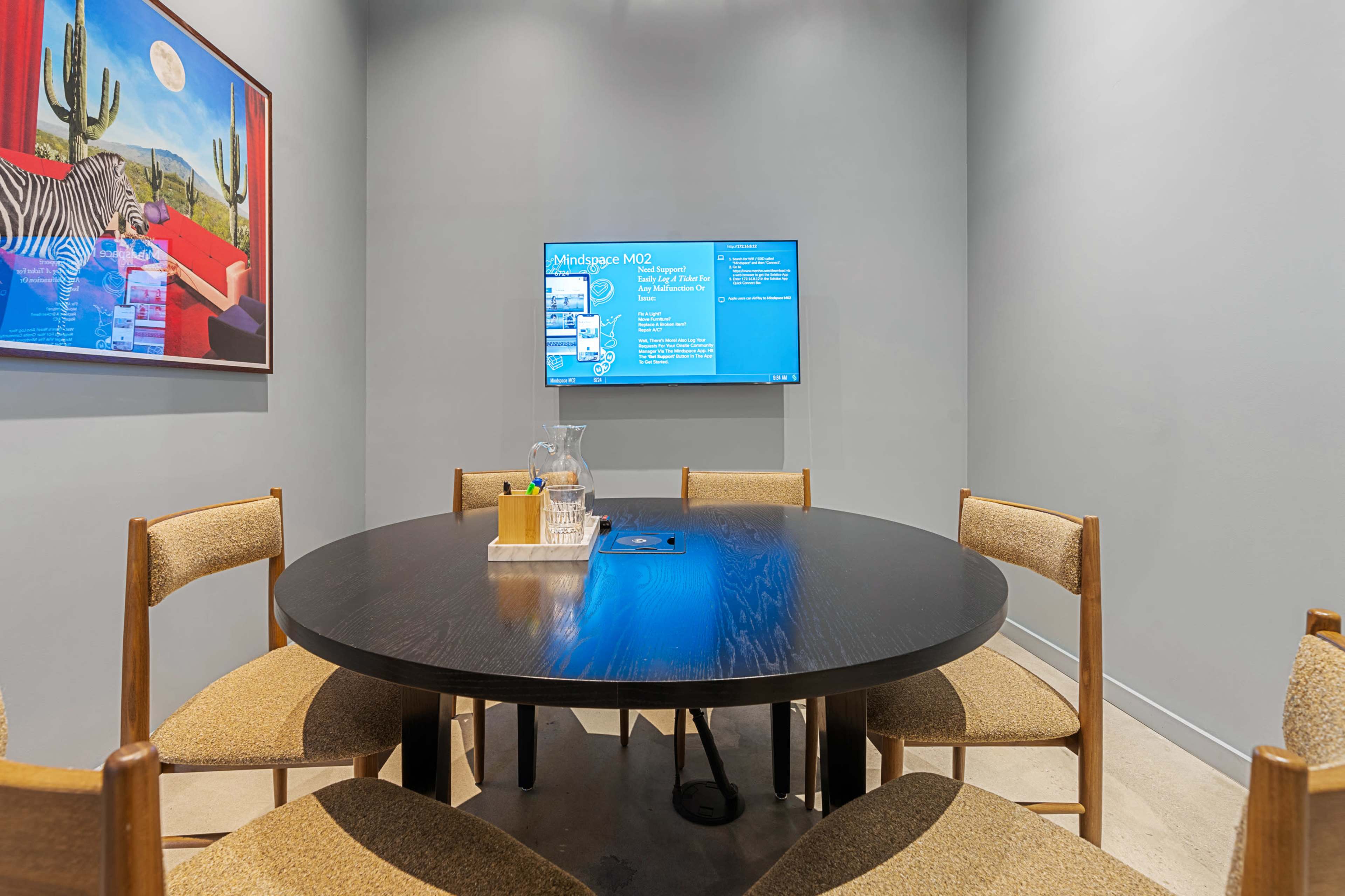 A round table with six chairs is set in a small conference room featuring a wall-mounted screen and artwork of a zebra.