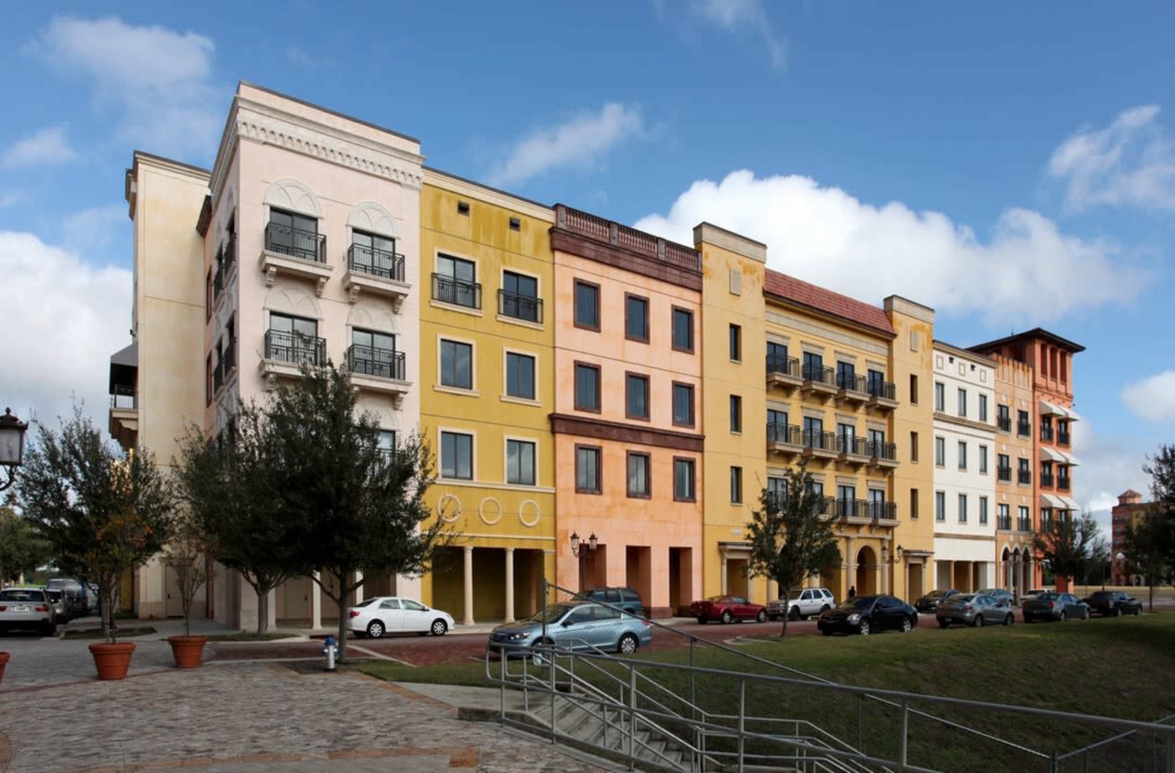 A row of multicolored buildings with balconies and windows along a street, alongside a grassy area.