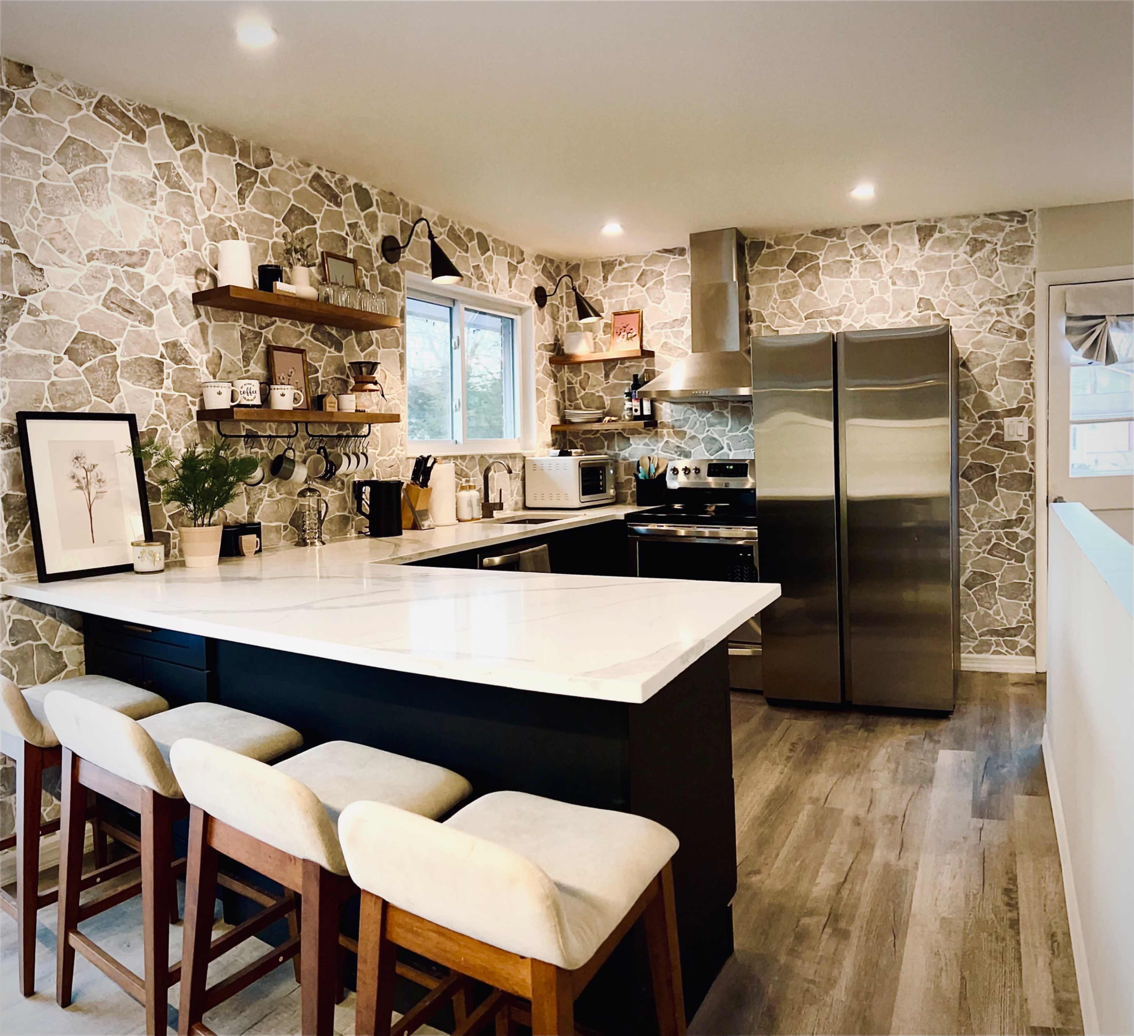 A modern kitchen features stone-patterned walls, a large island with seating, stainless steel appliances, and open shelving displaying kitchenware.