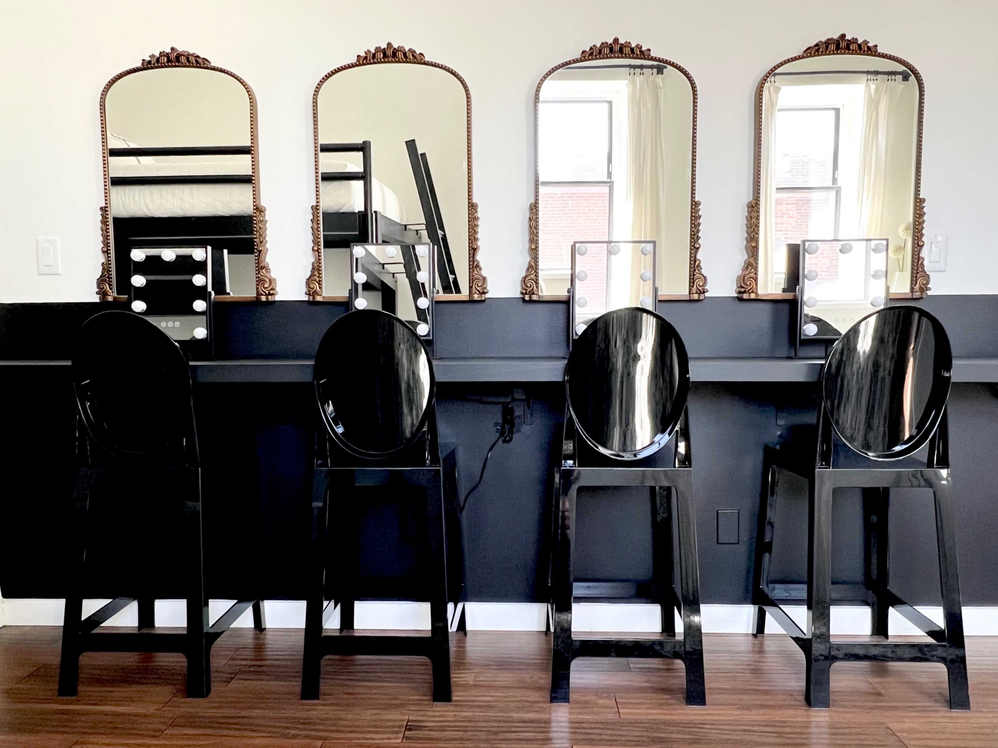 The image shows four black high stools positioned in front of a wall-mounted vanity with three ornate mirrors.