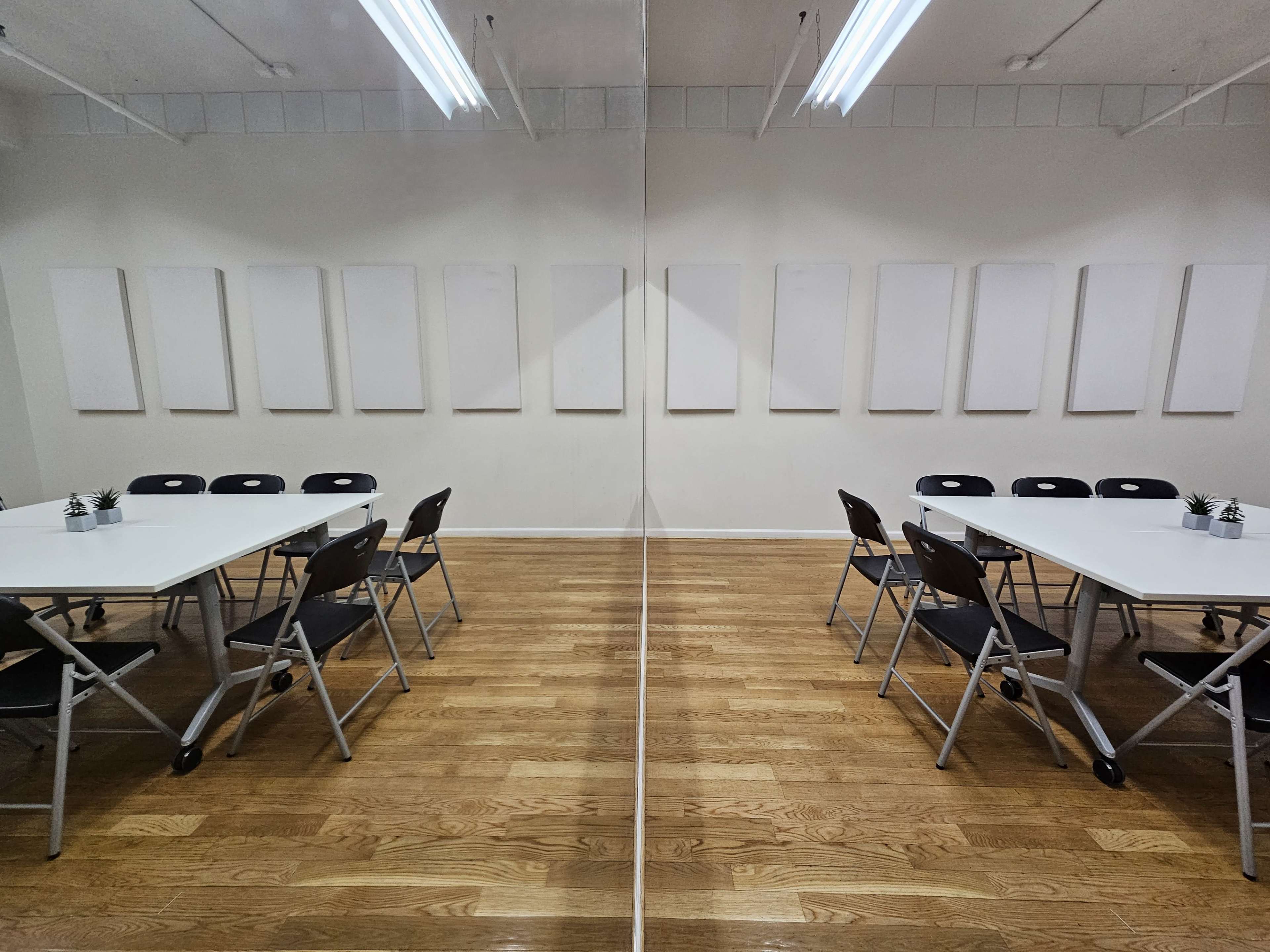 A bright meeting room features two tables with folding chairs arranged around them, facing a wall adorned with blank canvases.