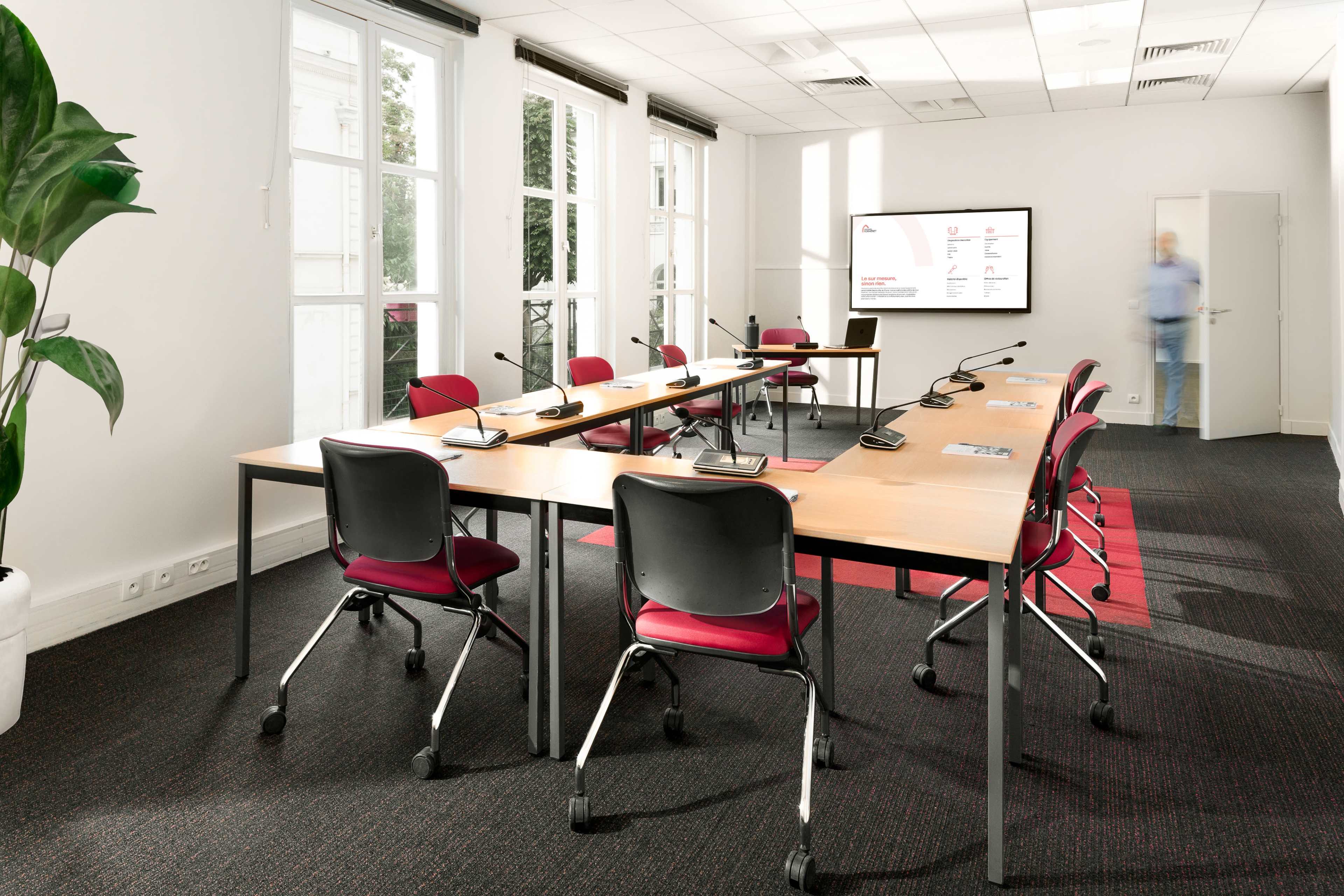 A modern conference room features a large table surrounded by red chairs, with a display screen on the wall and large windows allowing natural light.