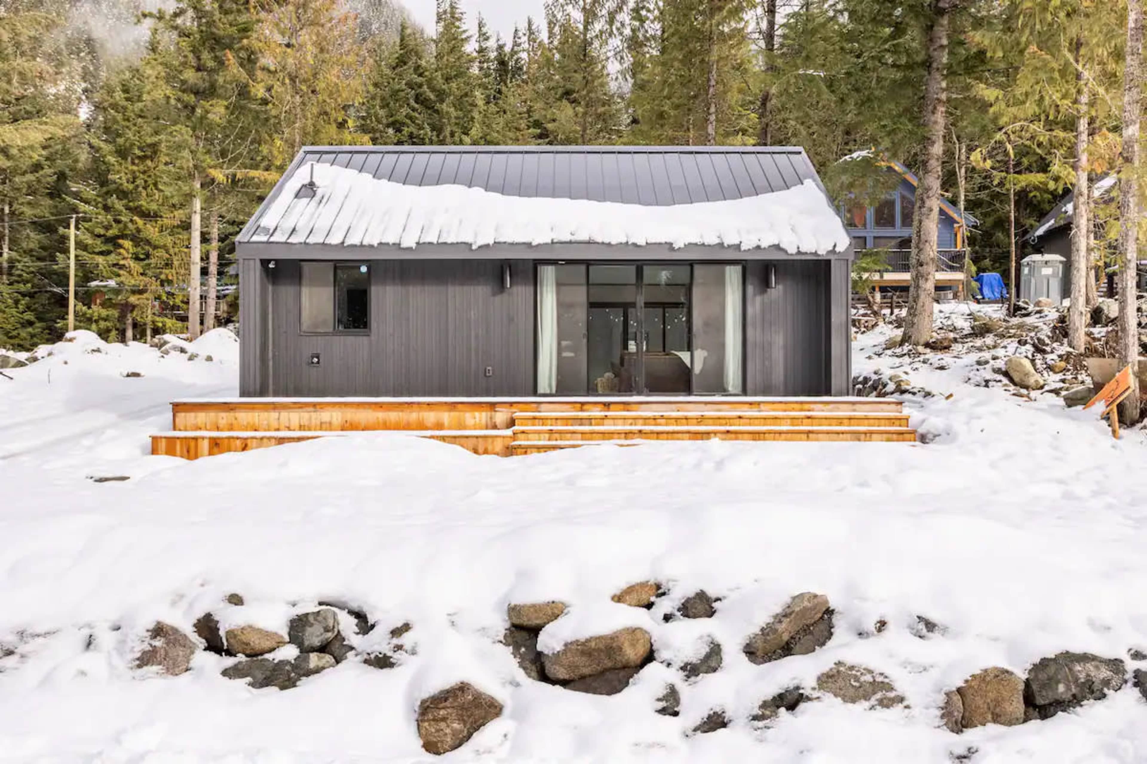 A modern cabin with a metal roof sits among snow-covered rocks and surrounding evergreen trees.