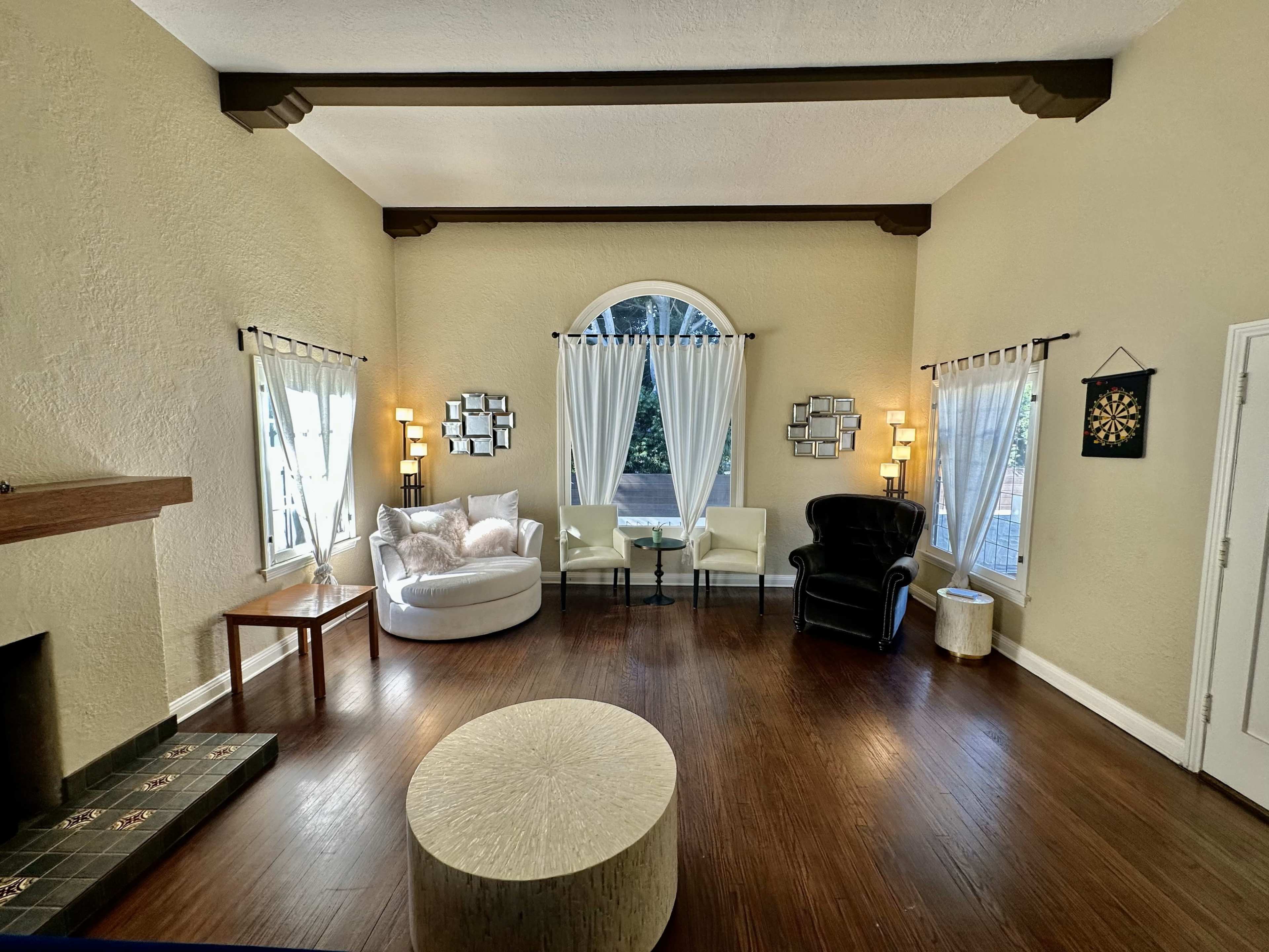 The image shows a spacious living room with a curved window, two armchairs, a round ottoman, and a fireplace.