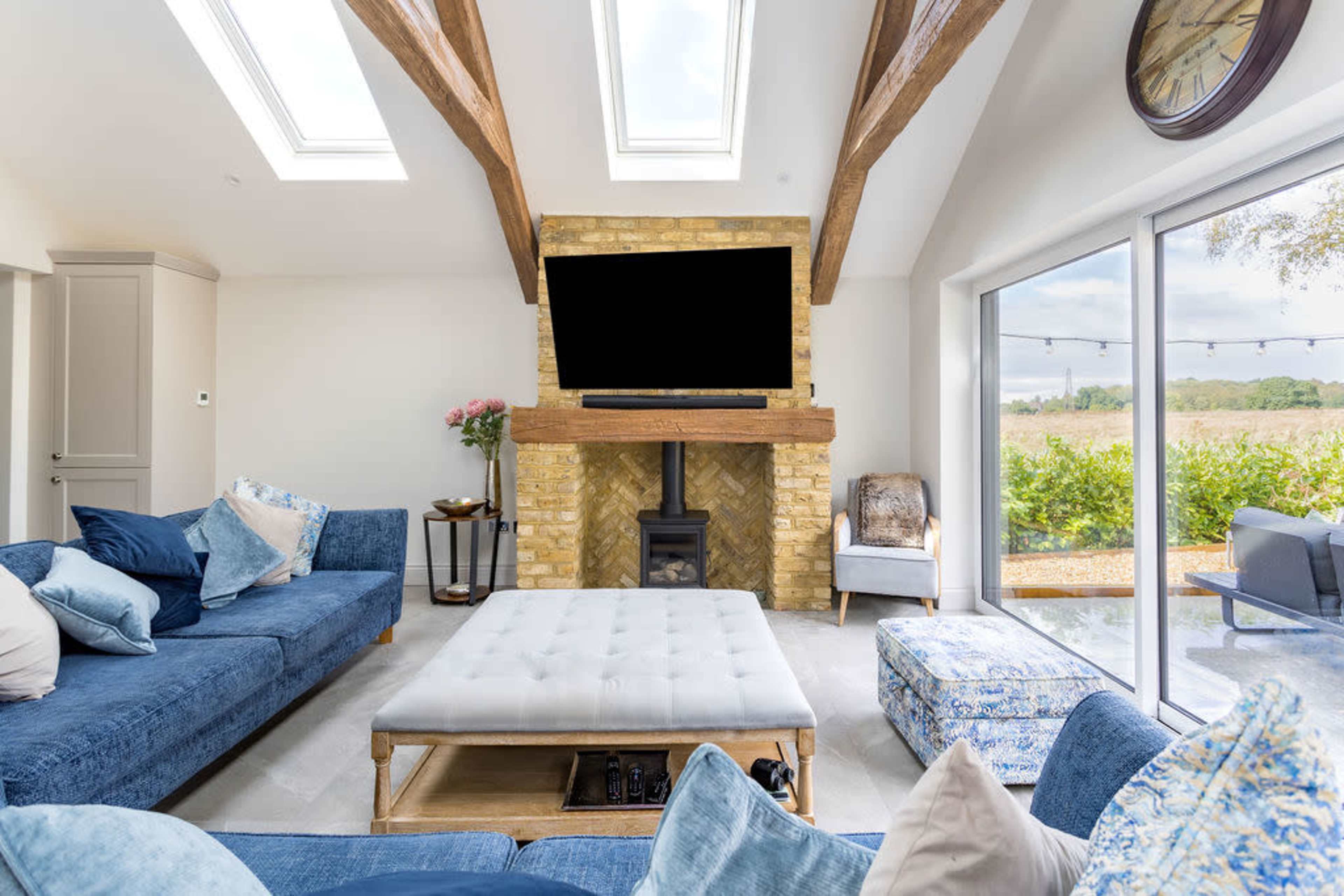 A stylish living room featuring a large blue sectional sofa, a light-colored ottoman in the center, and a stone fireplace with a flat-screen TV above it, all set against large windows that overlook greenery.