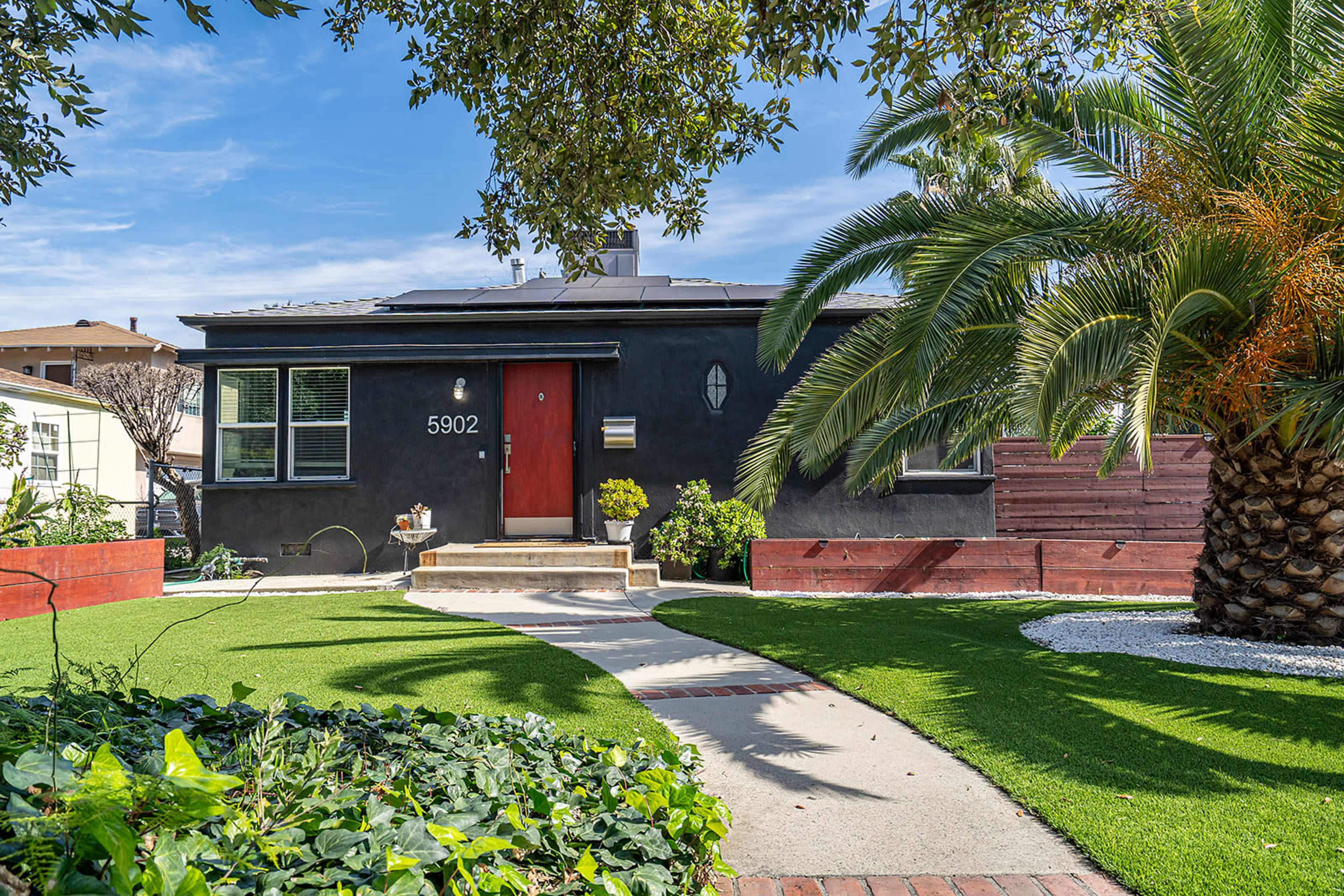 A black house with a red door is surrounded by lush green grass, palm trees, and garden beds.