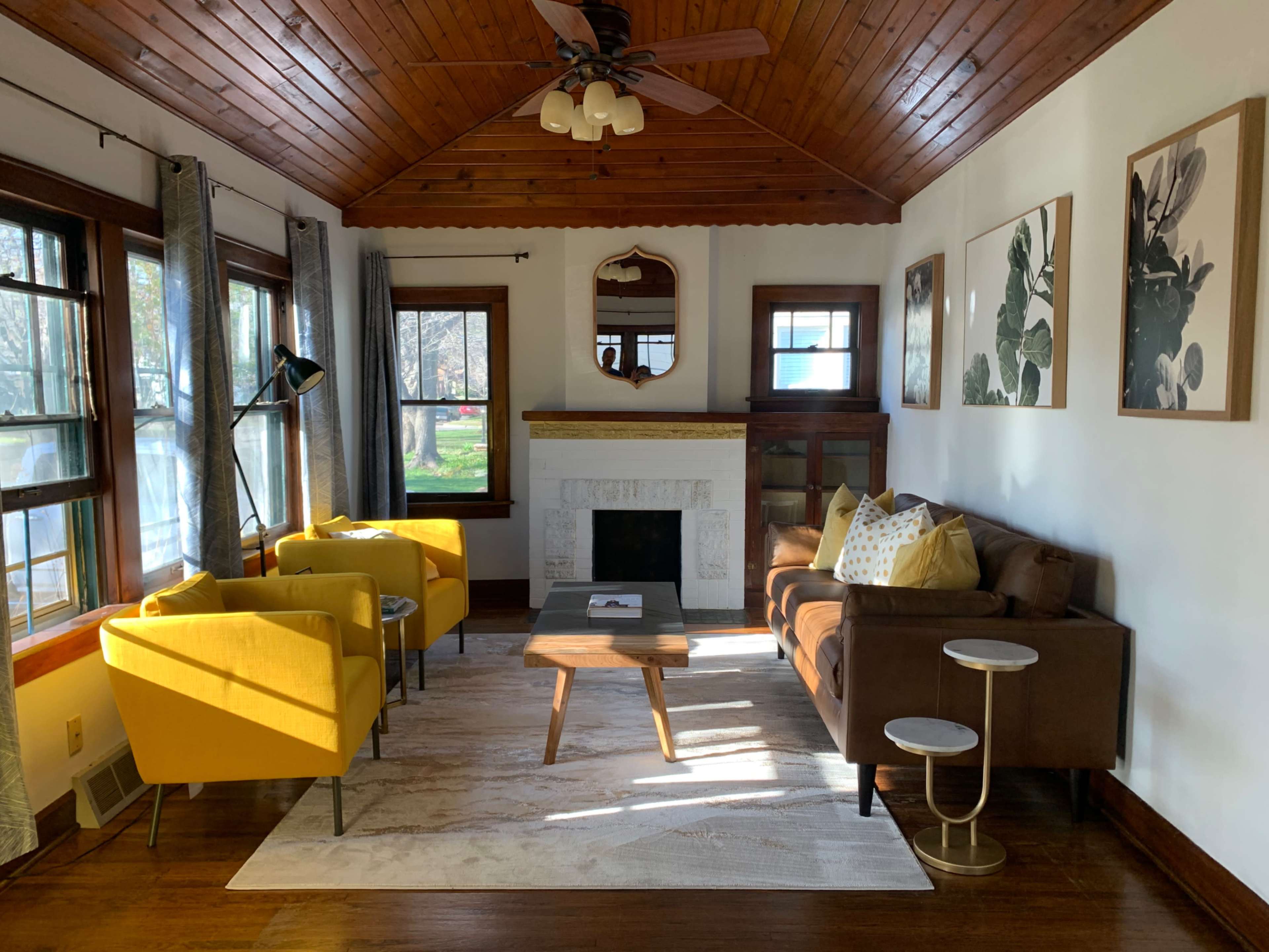 A living room with a wooden ceiling, a fireplace, two yellow armchairs, a brown sofa, and a coffee table placed on a light rug.