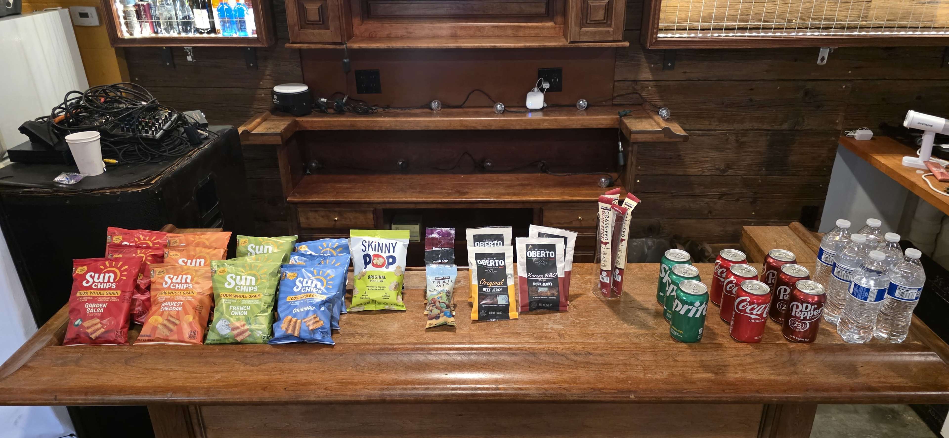 The image features a wooden table displaying various snack items, including bags of chips, granola bars, drinks, and bottled water, arranged neatly in rows.