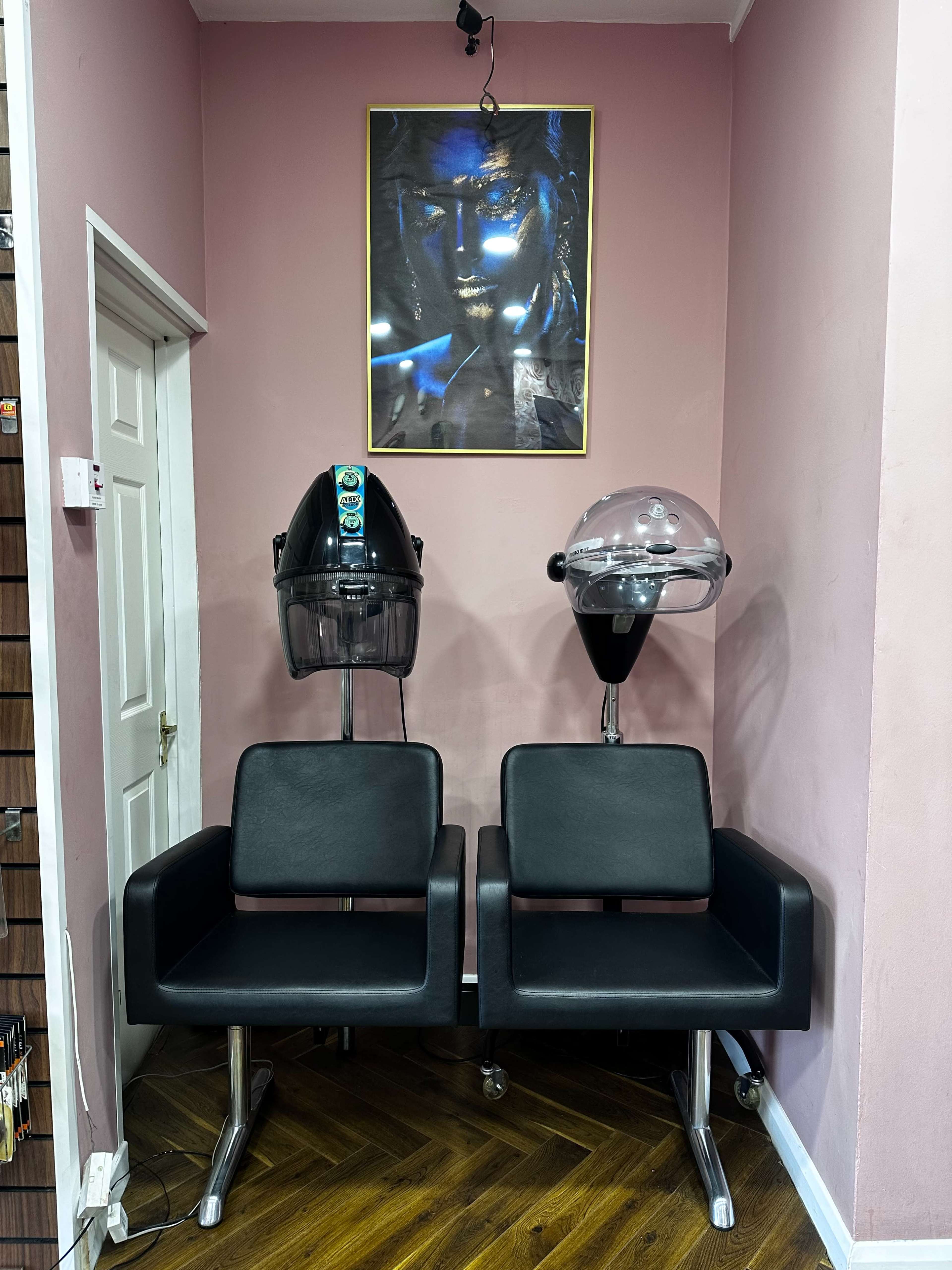 The image shows two black salon chairs positioned against a pink wall, with a framed artwork above and two hair dryers on stands nearby.
