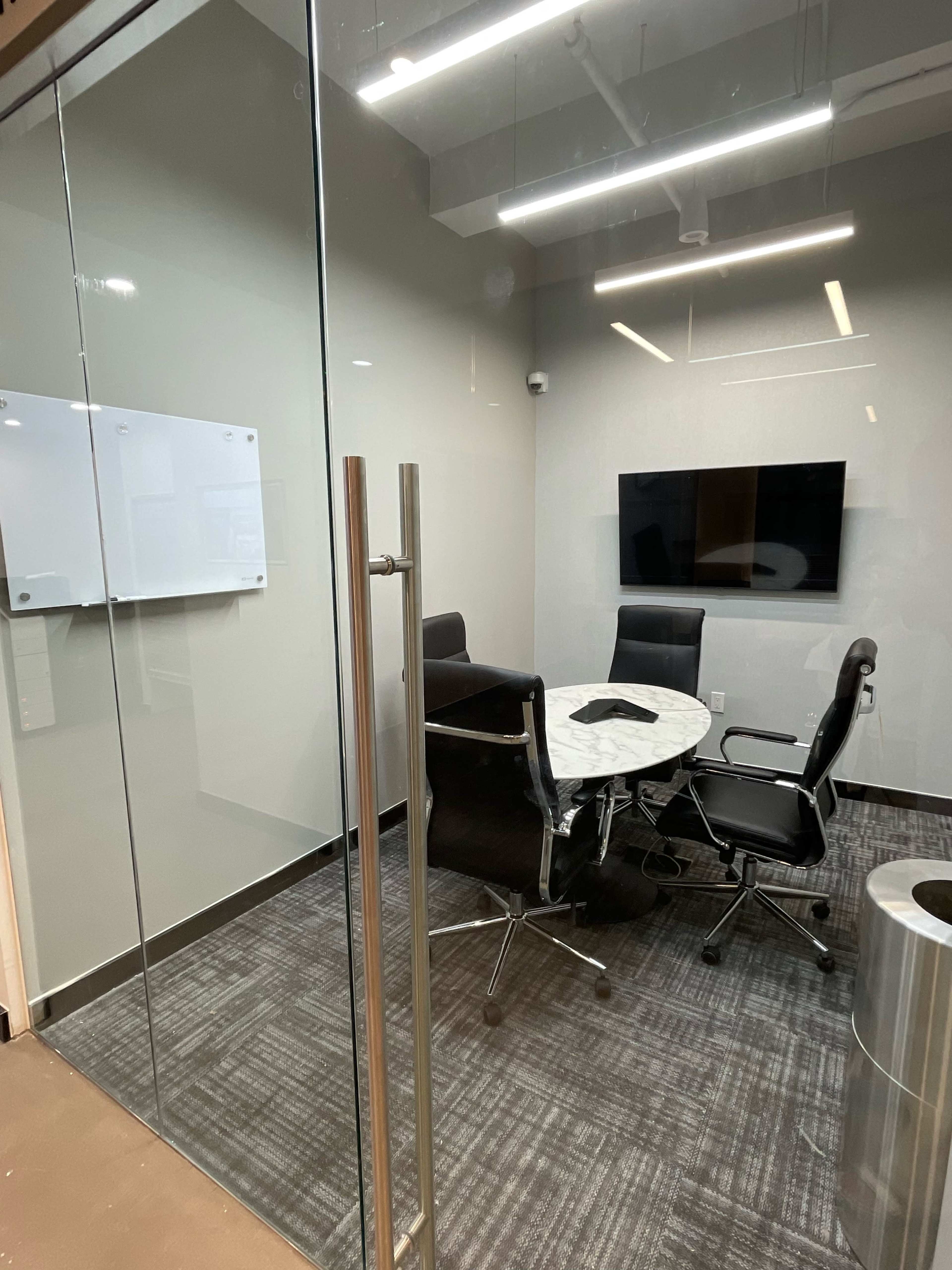 A modern conference room with a glass door, a round marble table, and four black office chairs.