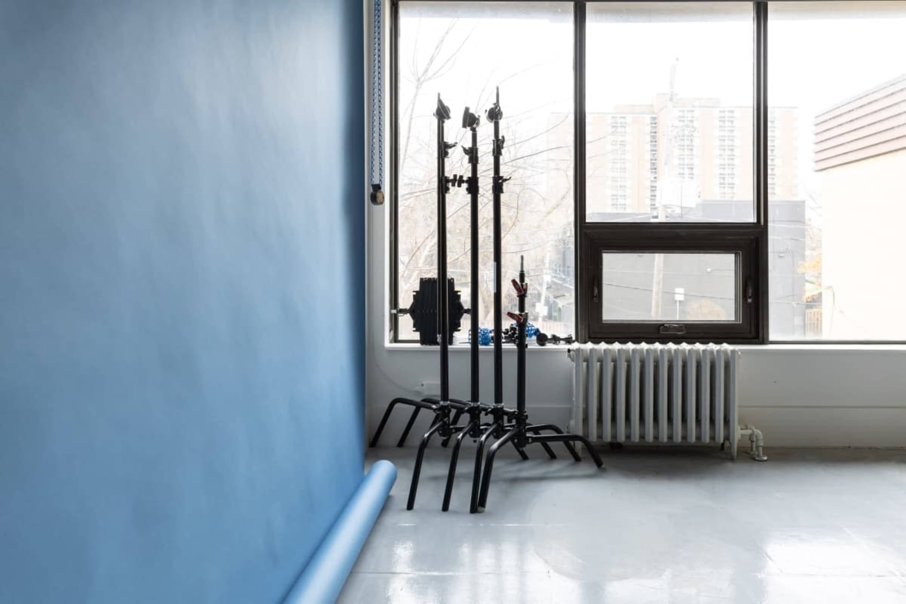 The image shows a minimalist interior with a blue wall, a window revealing a cityscape, and several black stands against the wall near a white radiator.