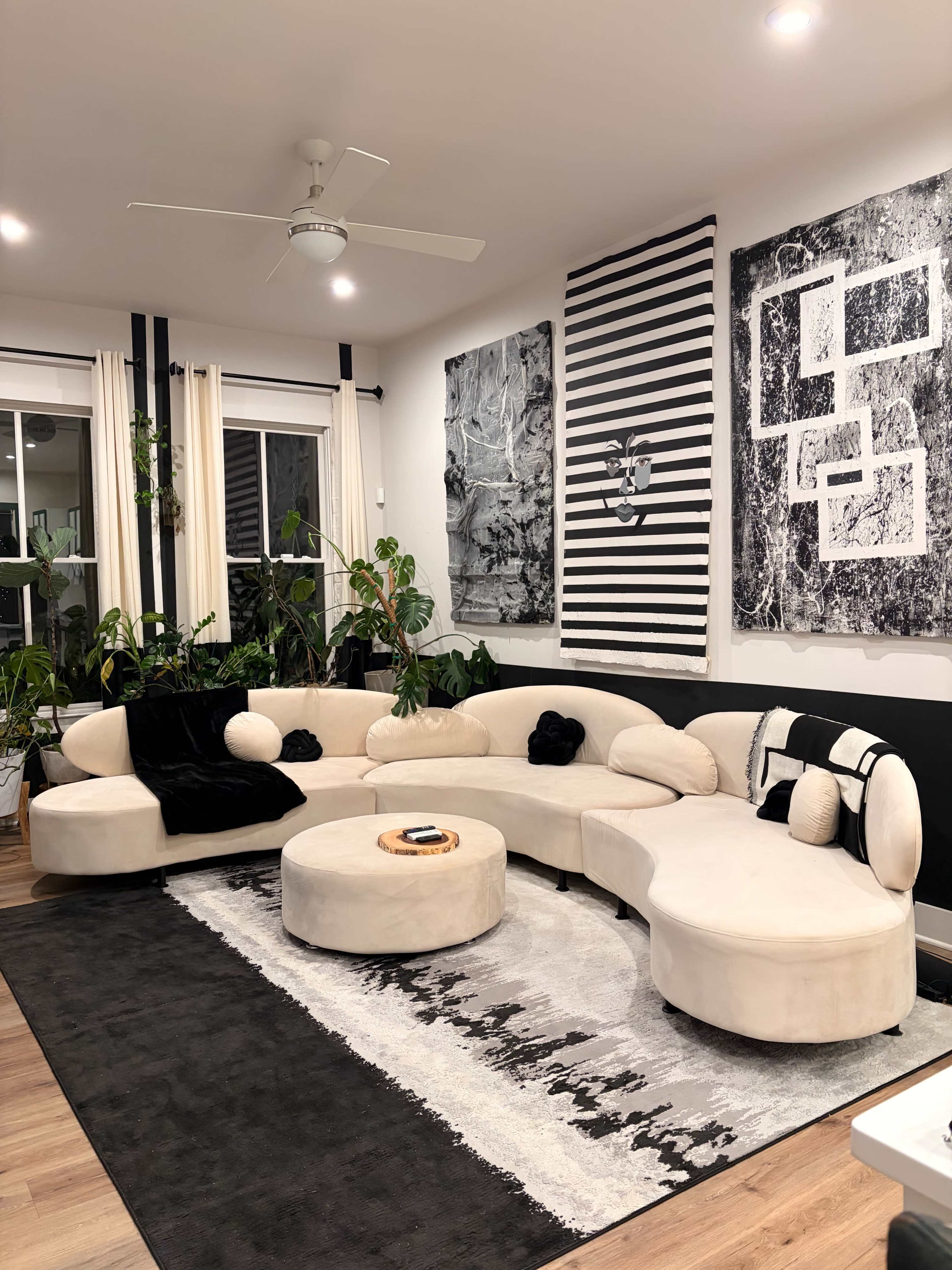 A modern living room features a curved beige sofa and a circular coffee table, surrounded by plants and adorned with abstract wall art.