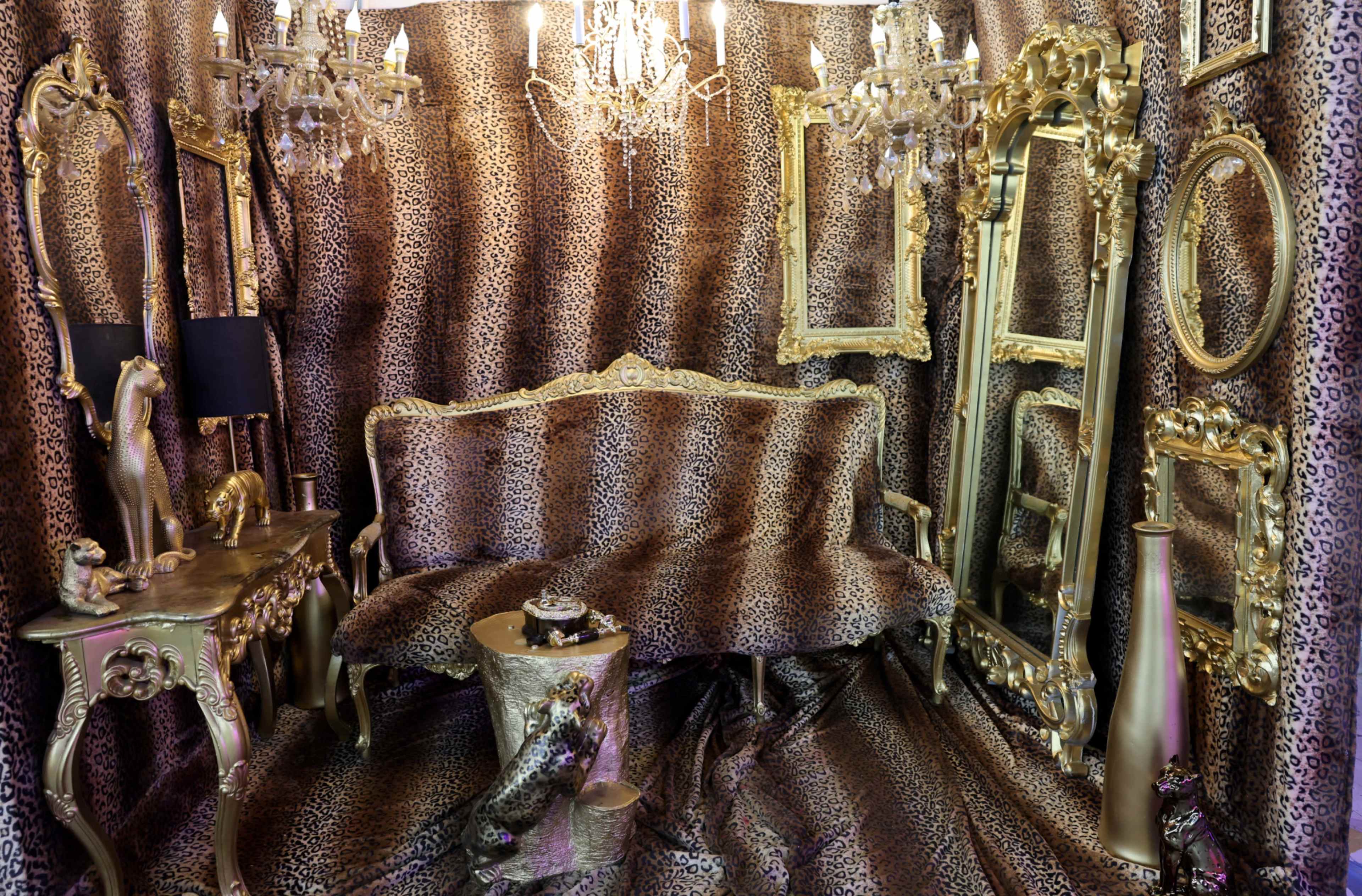 The image shows a lavish interior decorated with leopard print fabric, featuring gold-framed mirrors, a chandelier, and ornate furniture.