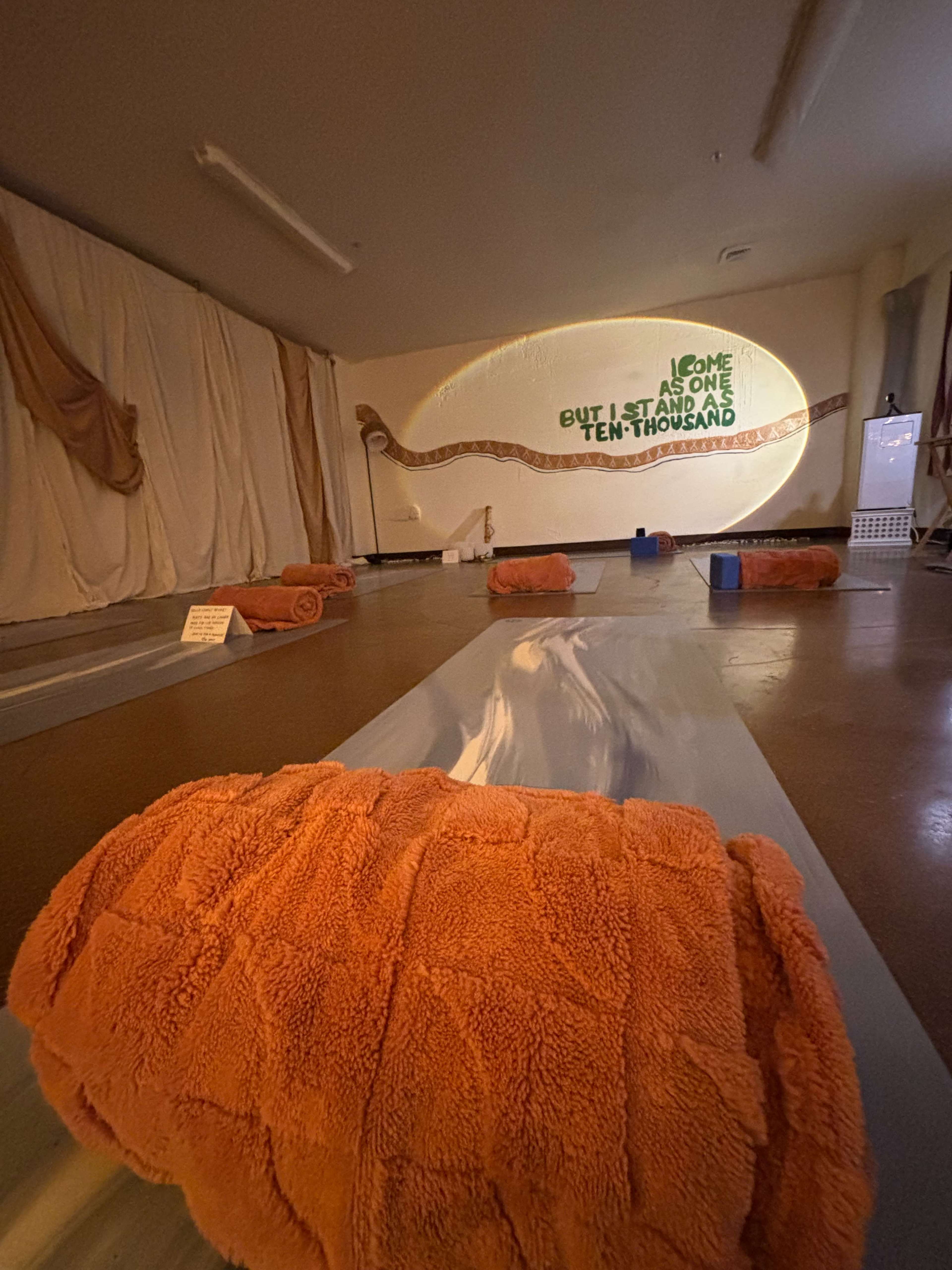 The image shows a meditation or yoga studio with rolled orange blankets on mats and a wall adorned with green text that reads, "Welcome, as one but I stand as ten thousand."