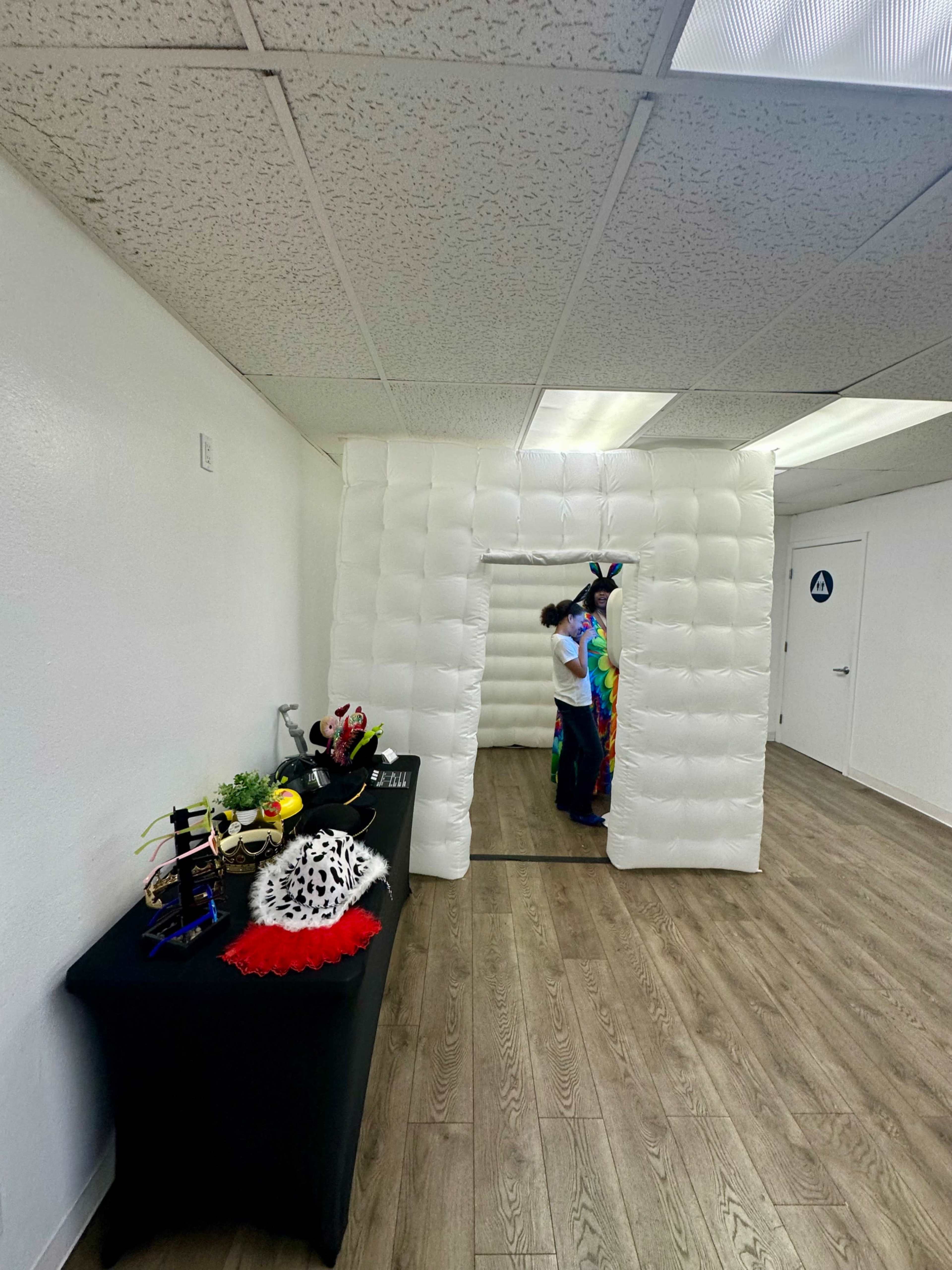 The image shows a white inflatable room with a person wearing a colorful outfit standing inside, and a table with various props placed nearby.