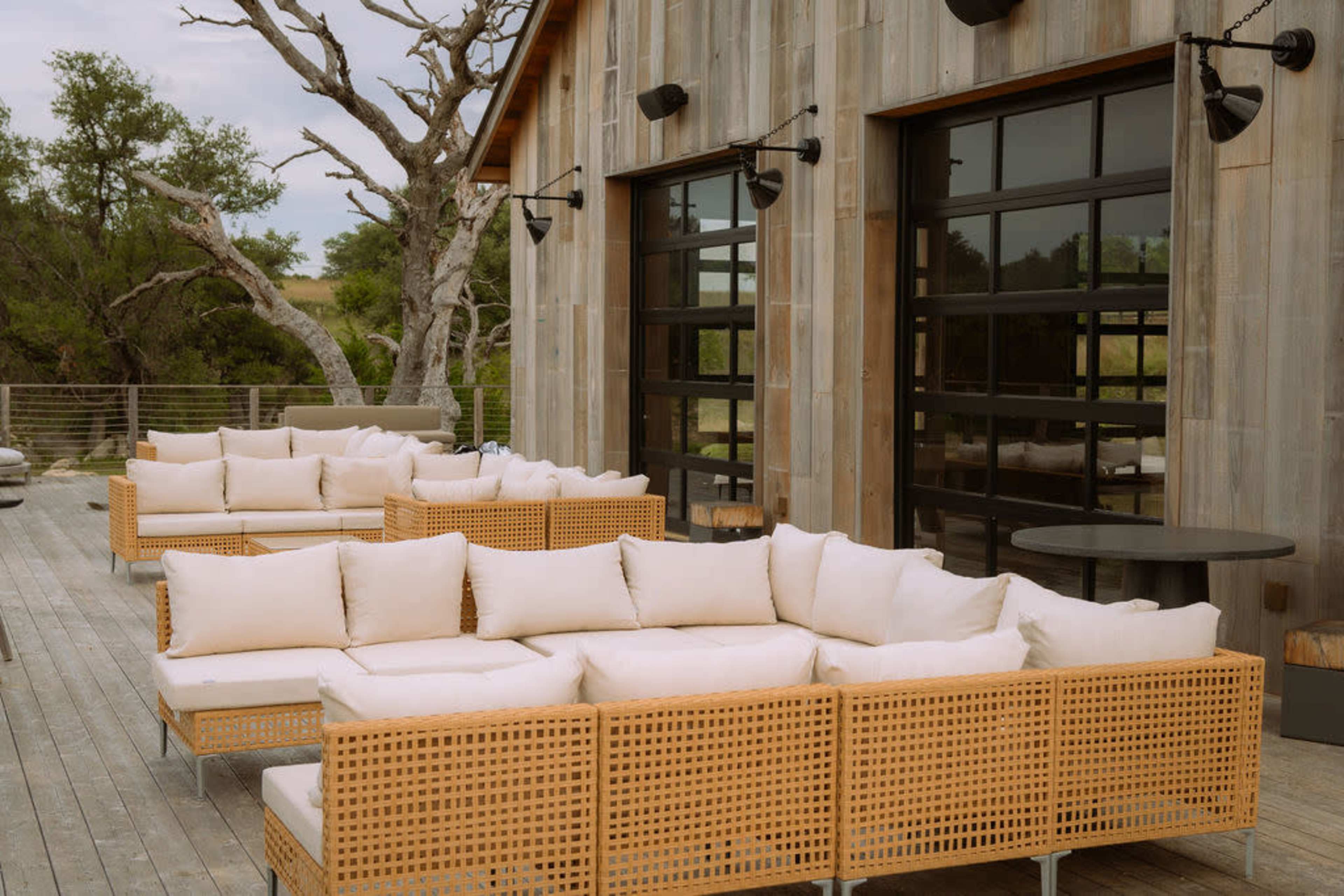 A wooden deck features several rattan seating arrangements with white cushions, adjacent to large glass doors of a modern building.
