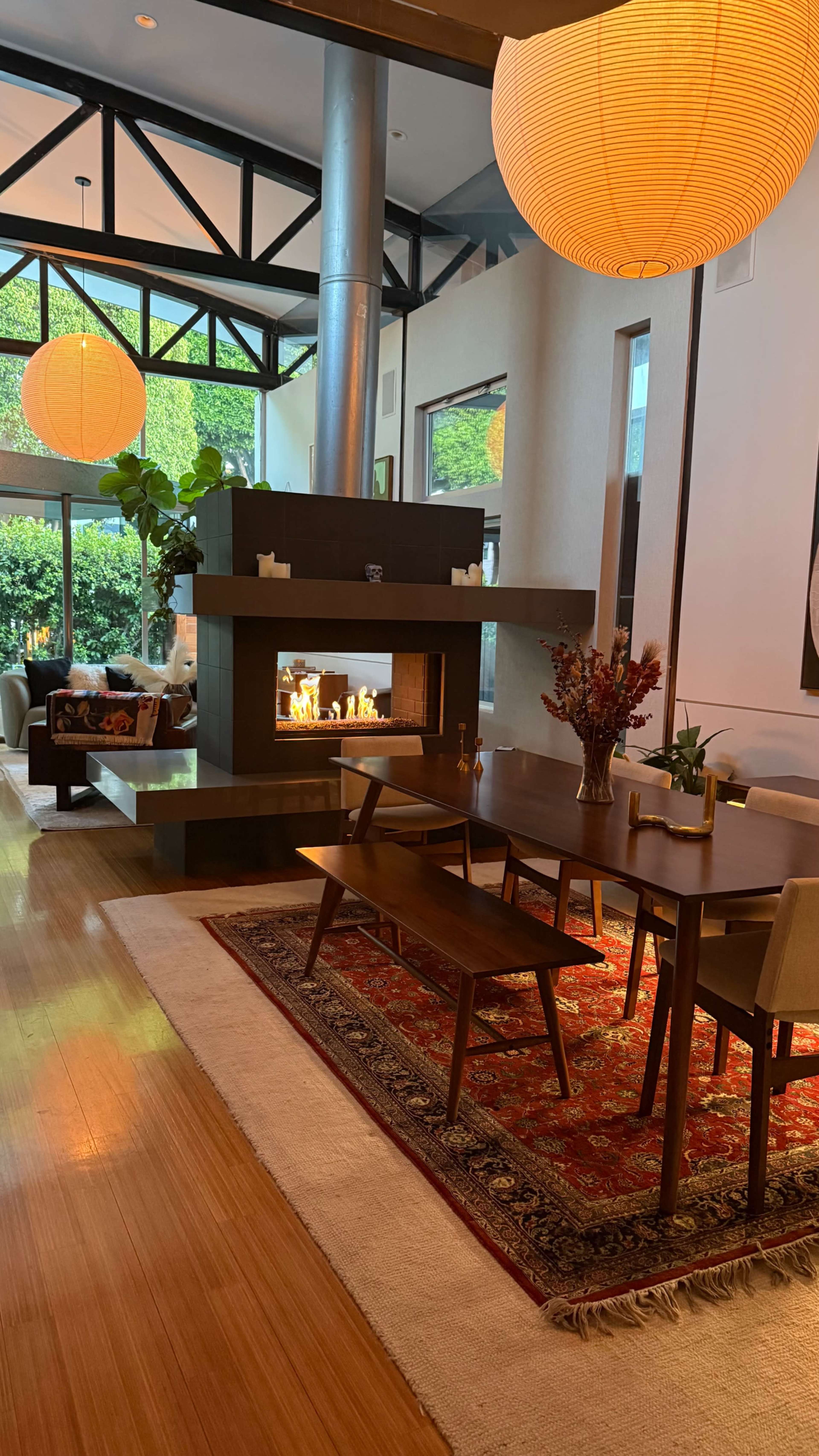 The image shows a modern living space with a dining area featuring a wooden table, surrounded by chairs, and a fireplace in the background, illuminated by large hanging lanterns.