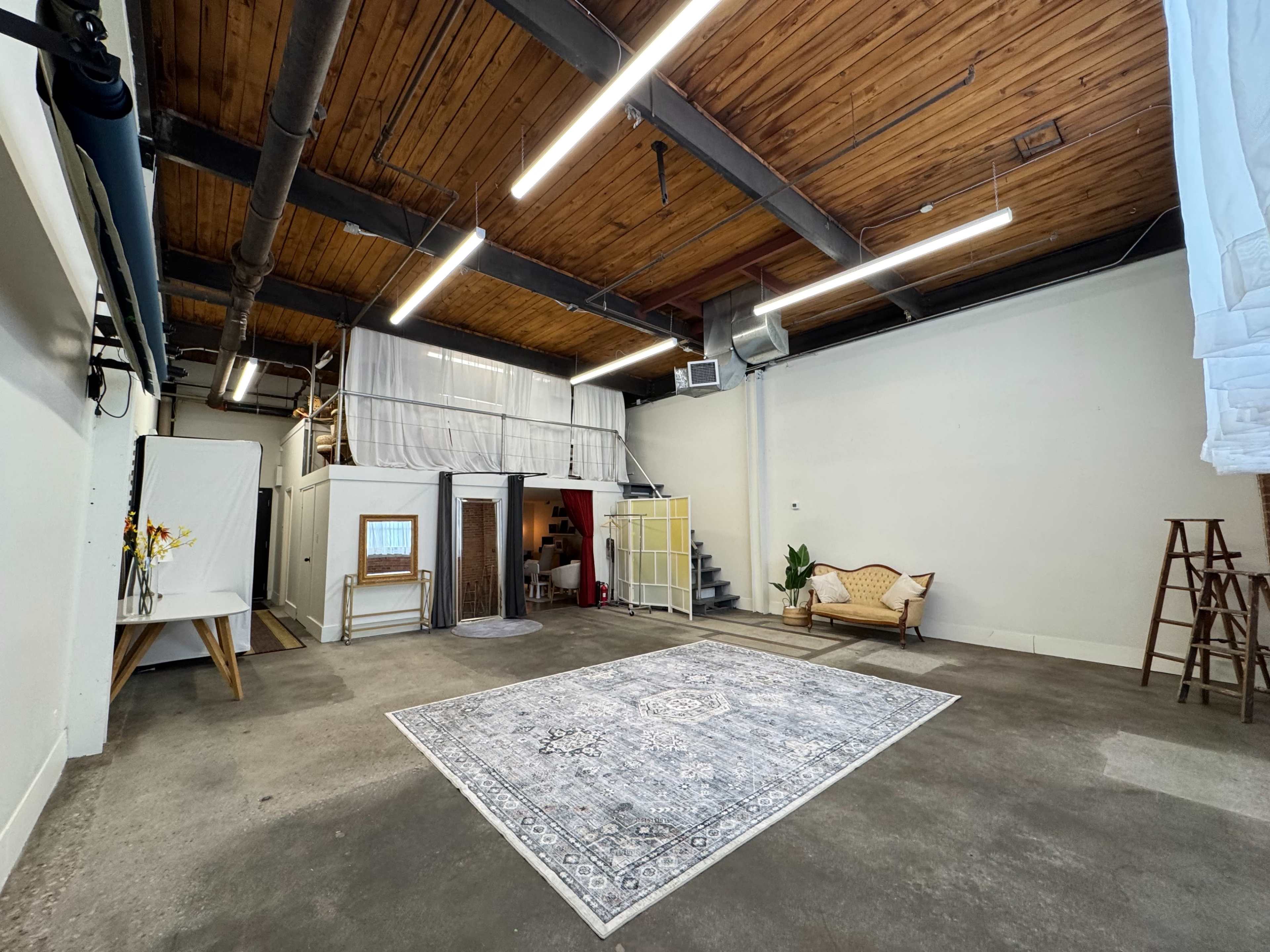 The image shows a spacious, industrial-style room with a wooden ceiling, concrete floor, and a large decorative rug in the center, along with a sofa and various photography equipment.