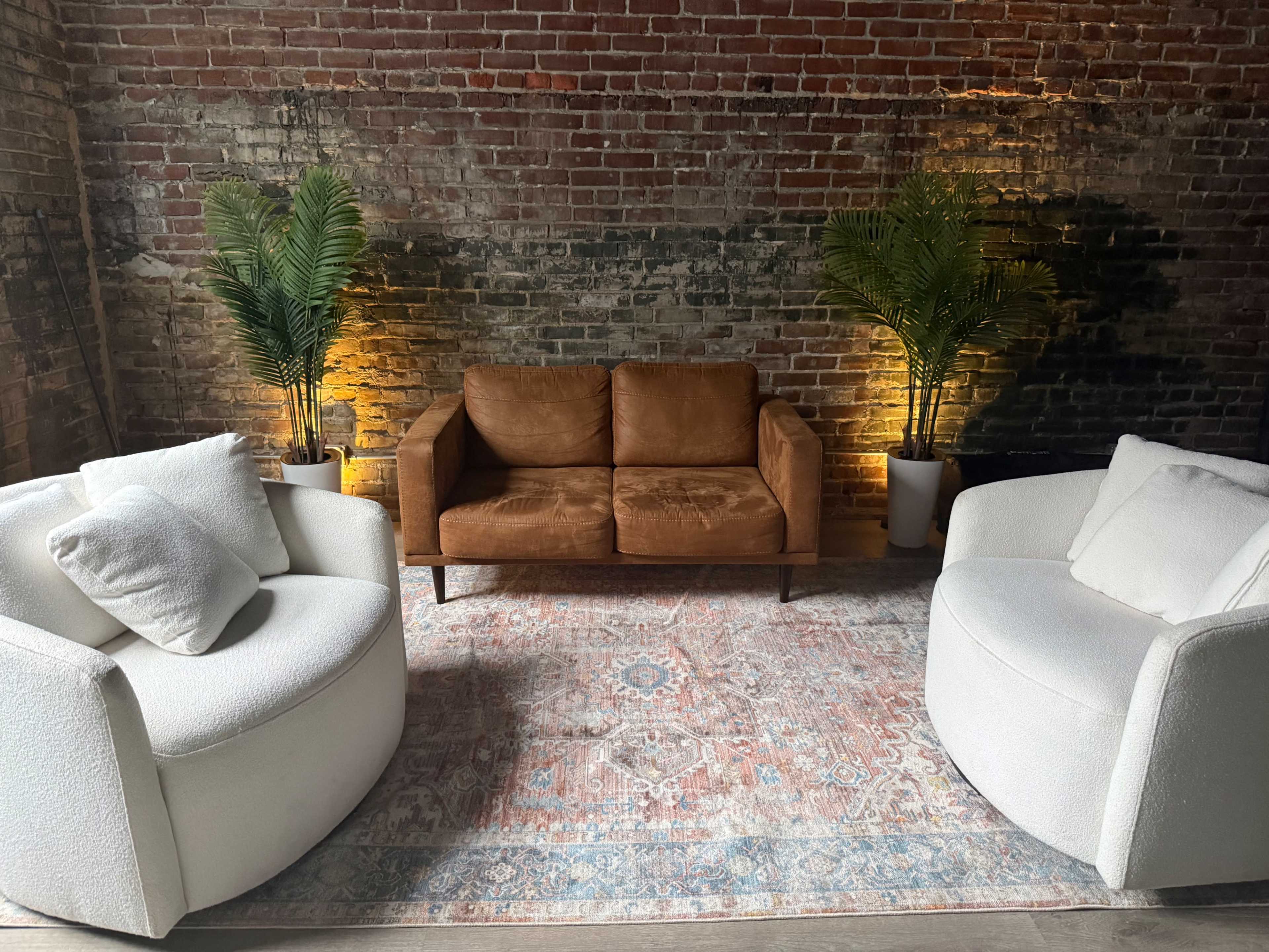 The image shows a cozy seating arrangement with a brown sofa and two white round chairs on a patterned rug in front of a brick wall, accented by indoor plants and soft lighting.