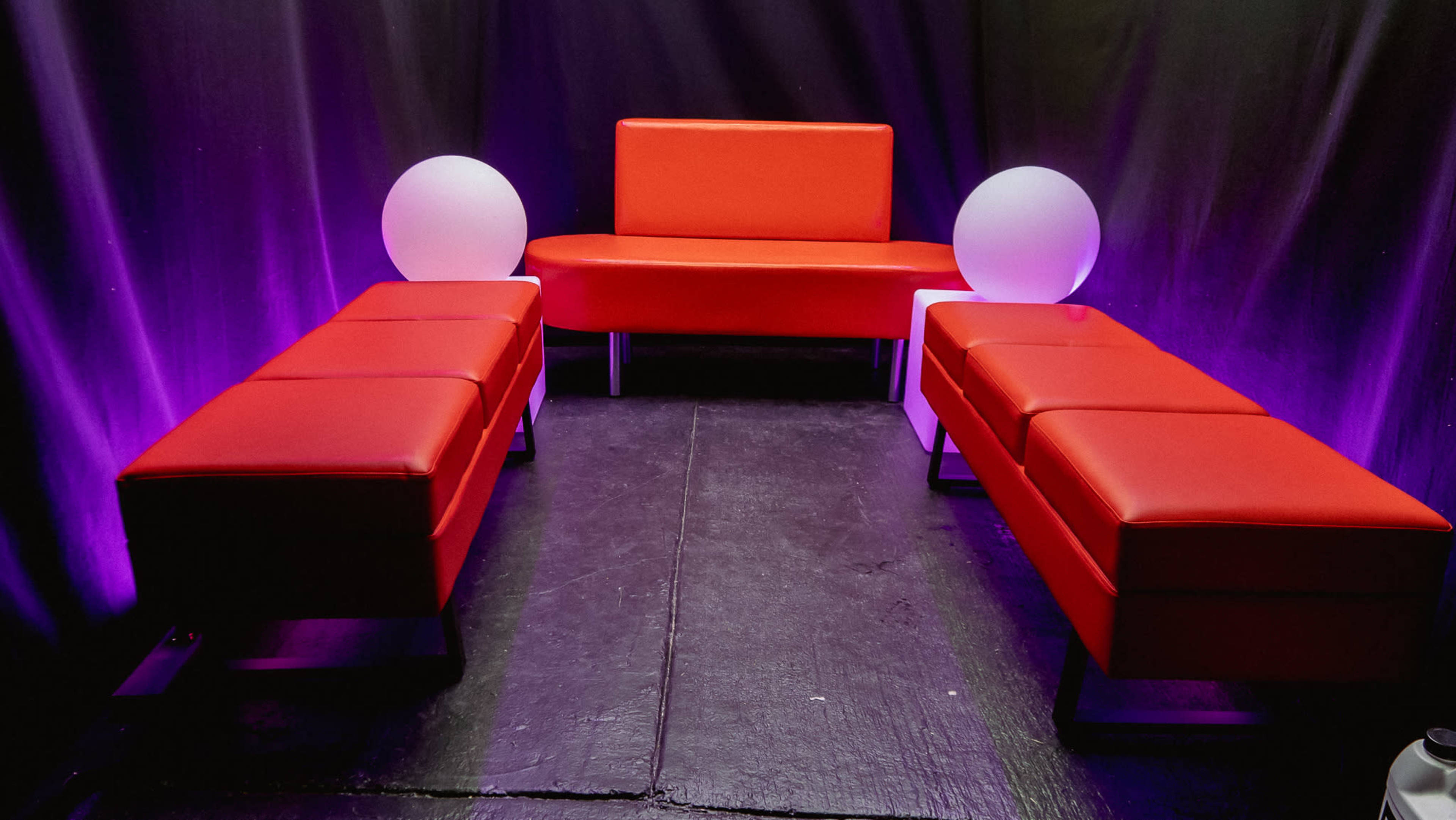 A small seating area with a red couch and two matching red benches flanking white spherical light fixtures, all set against a dark backdrop illuminated with purple light.