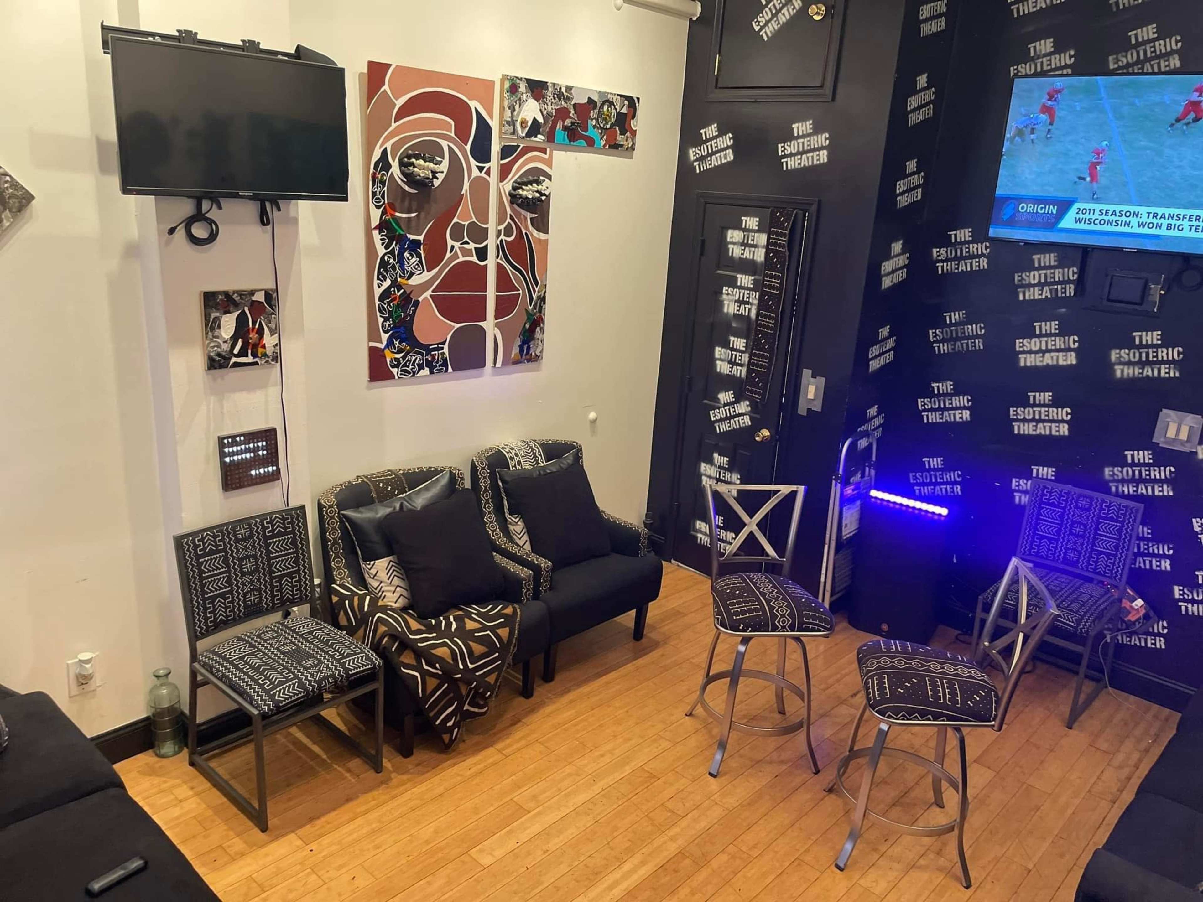 A small, modern room furnished with patterned chairs and a television on the wall, featuring bold artwork and decorative text on the walls.