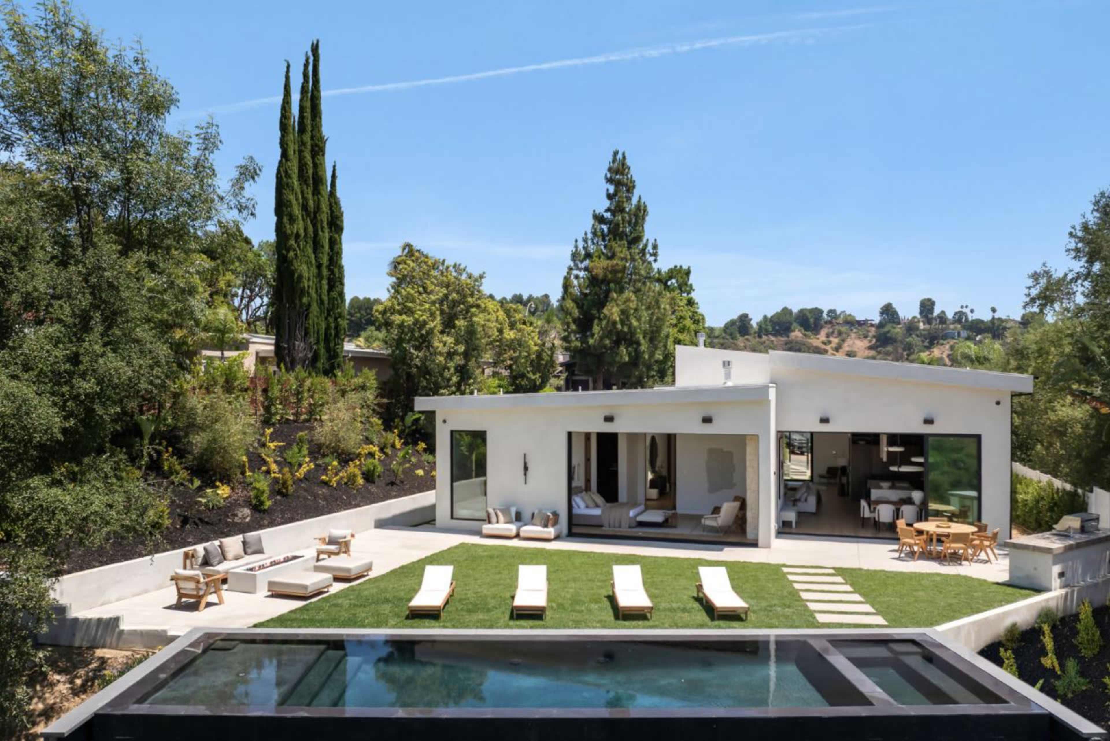 A modern white house with large windows overlooks a landscaped yard featuring a swimming pool and lounge chairs.