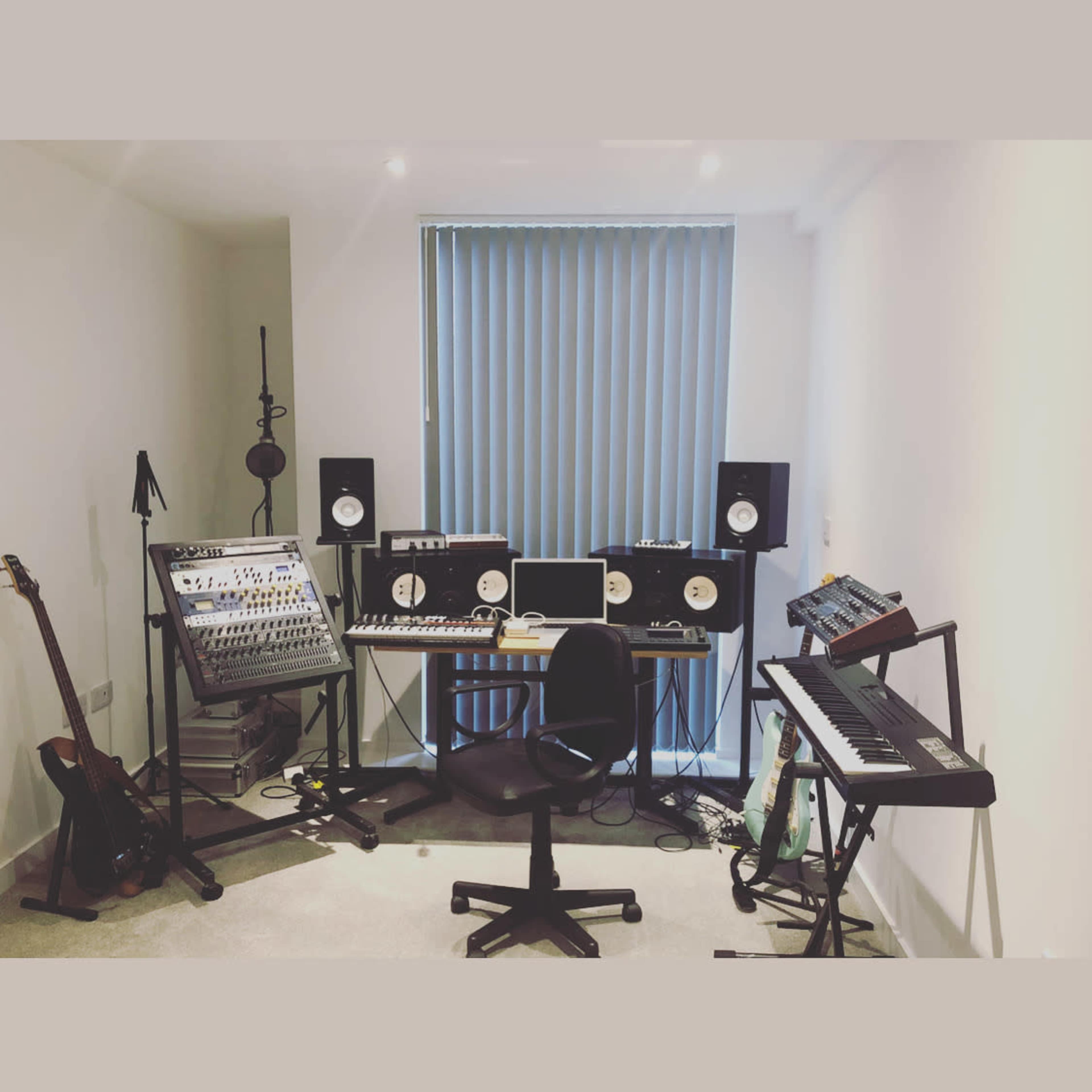 The image shows a music production studio with various instruments, a keyboard, audio monitors, and a computer setup in a room with vertical blinds.