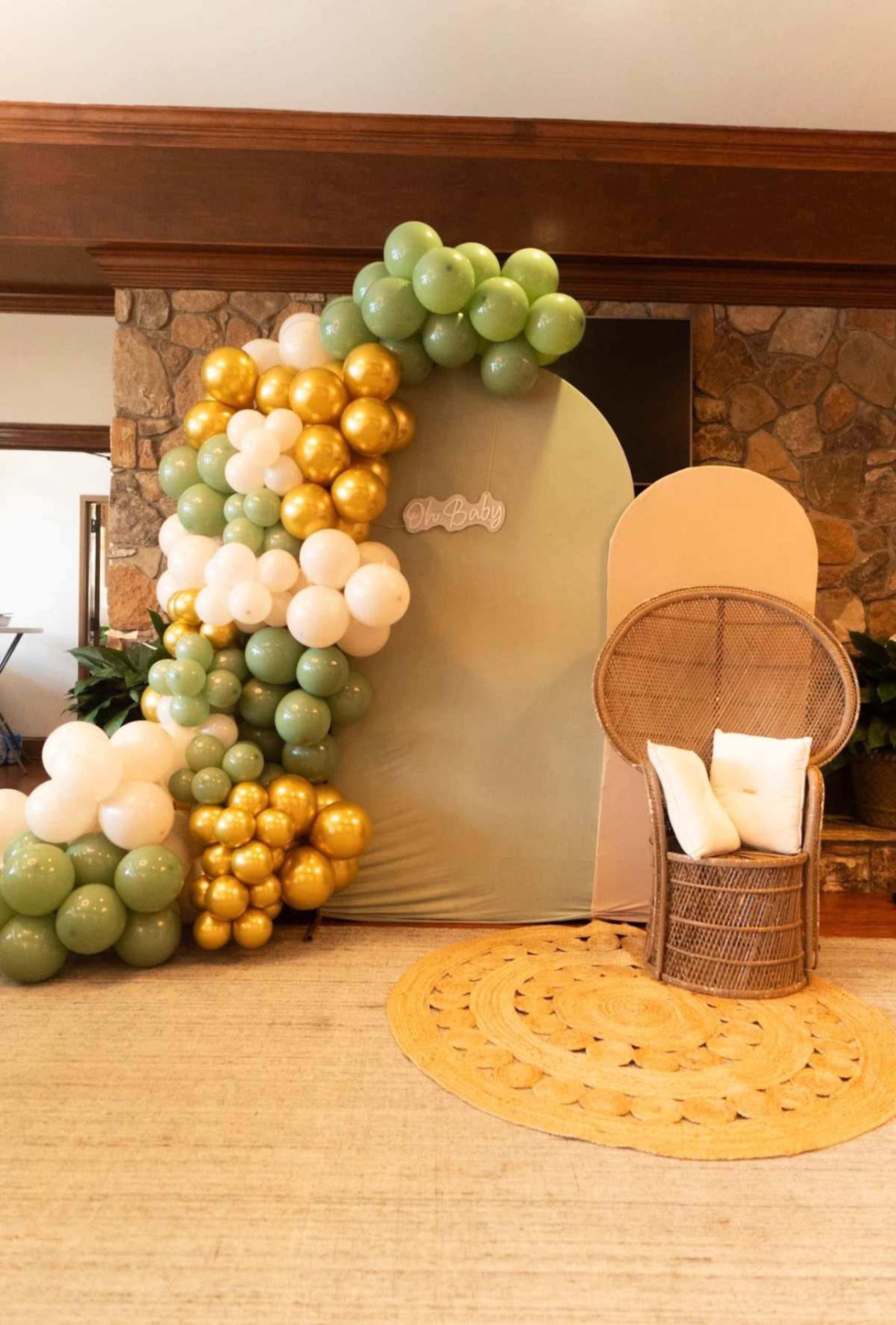 The scene features a decorative balloon arch in shades of green, white, and gold beside a wicker chair with two pillows on a circular, woven rug.