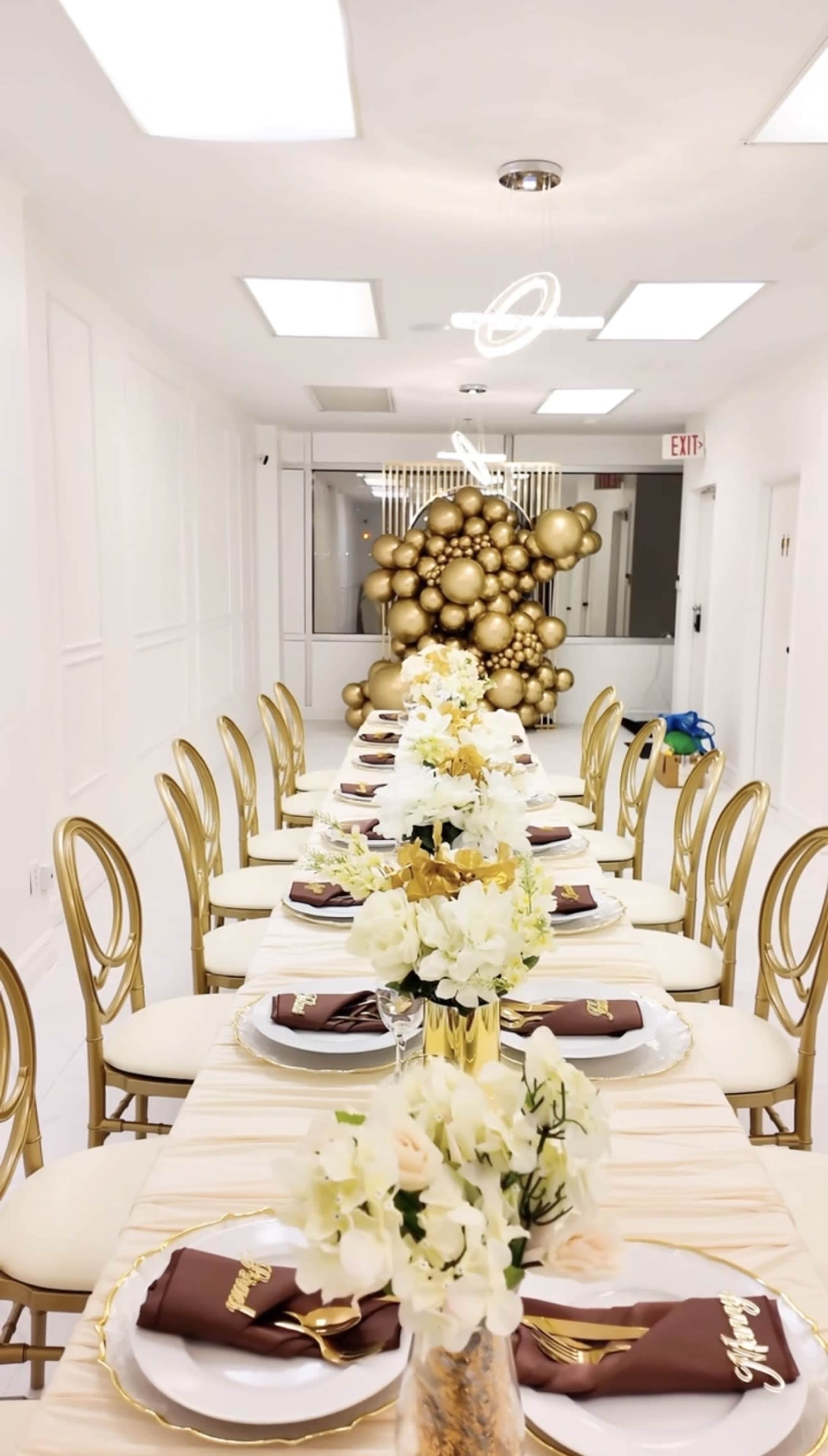 A long dining table is set with white plates and gold accents, flanked by gold chairs, and decorated with floral centerpieces and a large gold balloon display in the background.