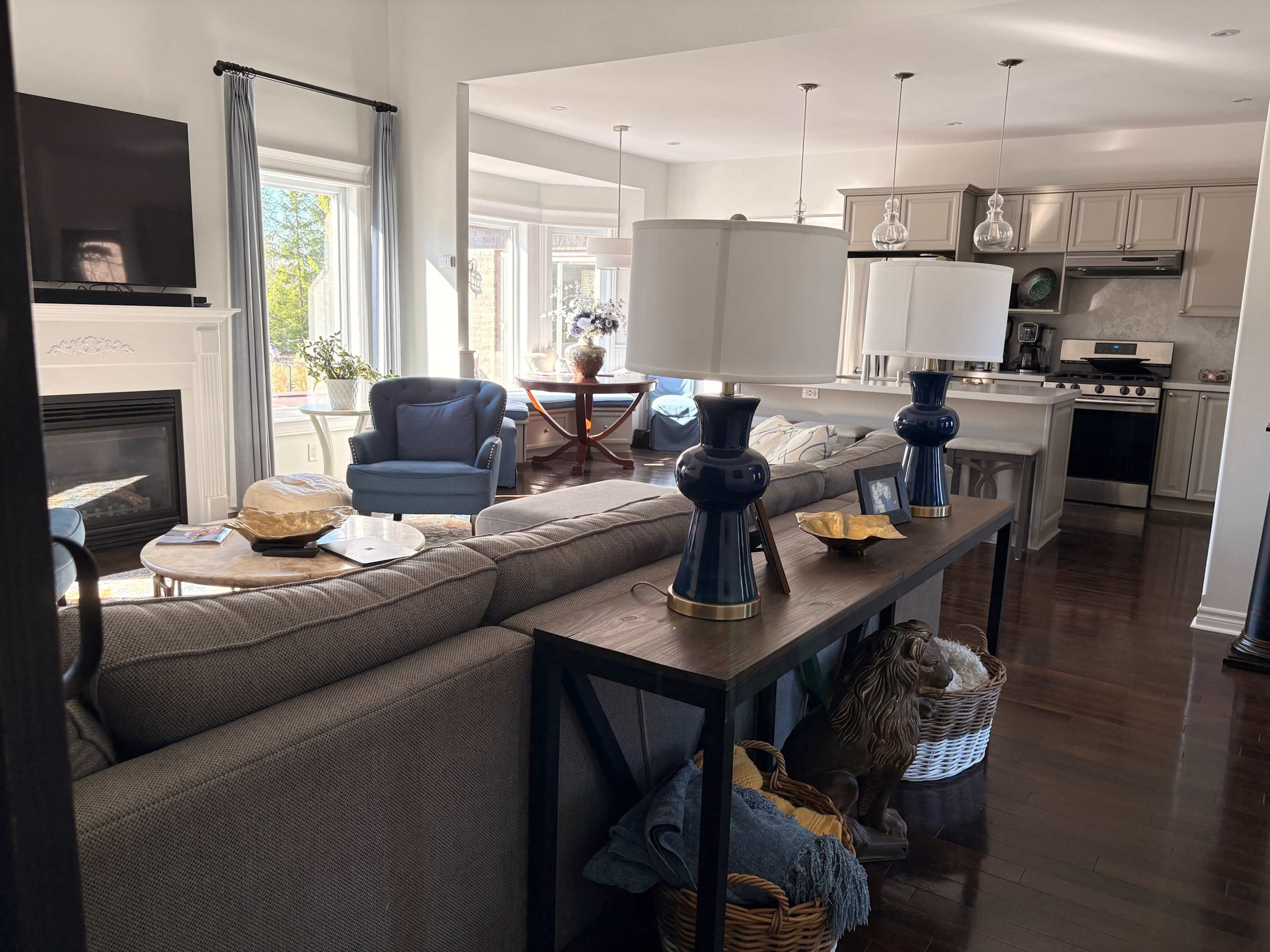 The image shows a bright living room featuring a sectional sofa, decorative lamps on a table, and a view of an adjacent kitchen.