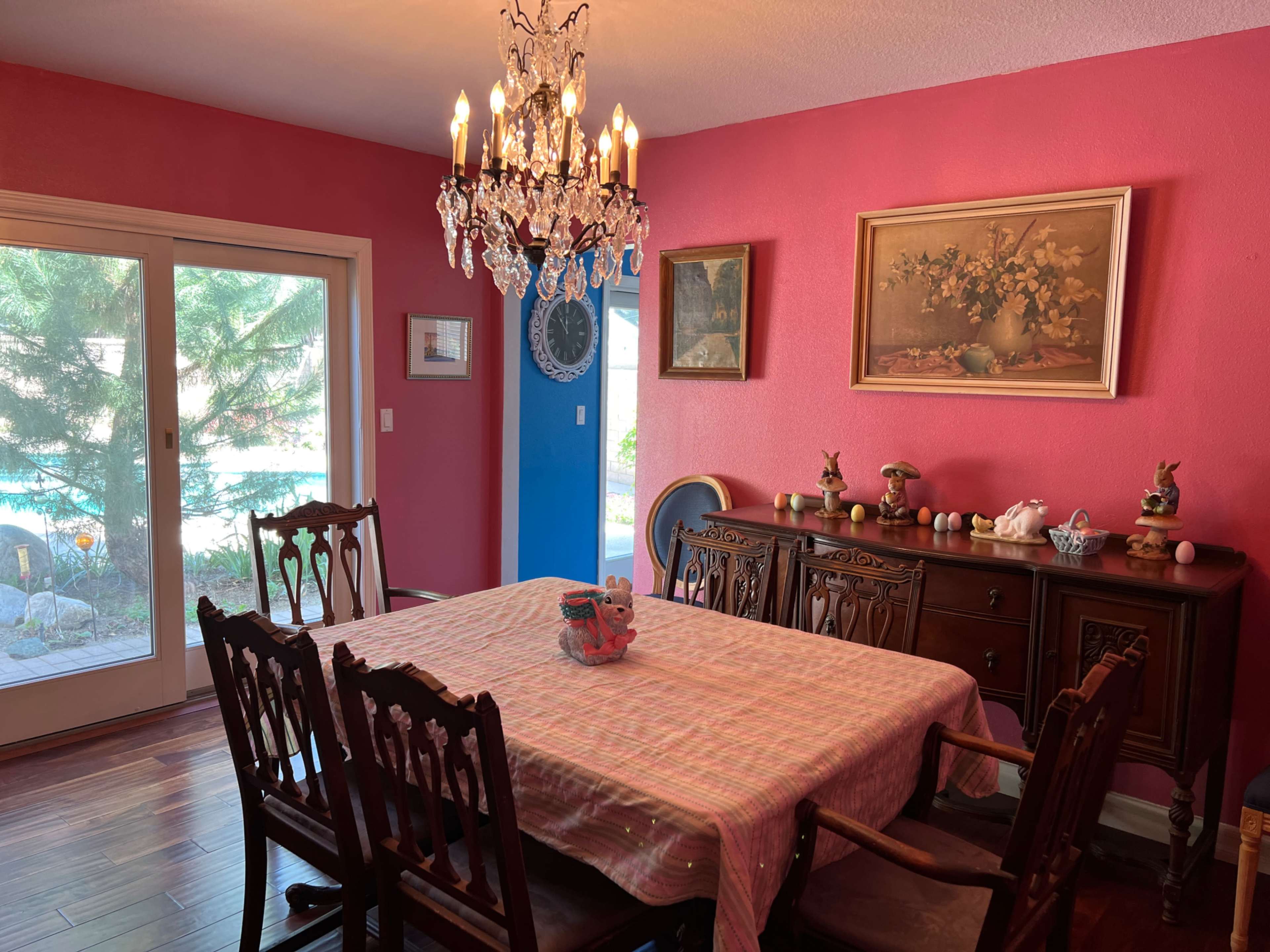 The dining room features a wooden table surrounded by chairs, a chandelier overhead, and a sideboard adorned with decorative items, all against a bright pink wall.
