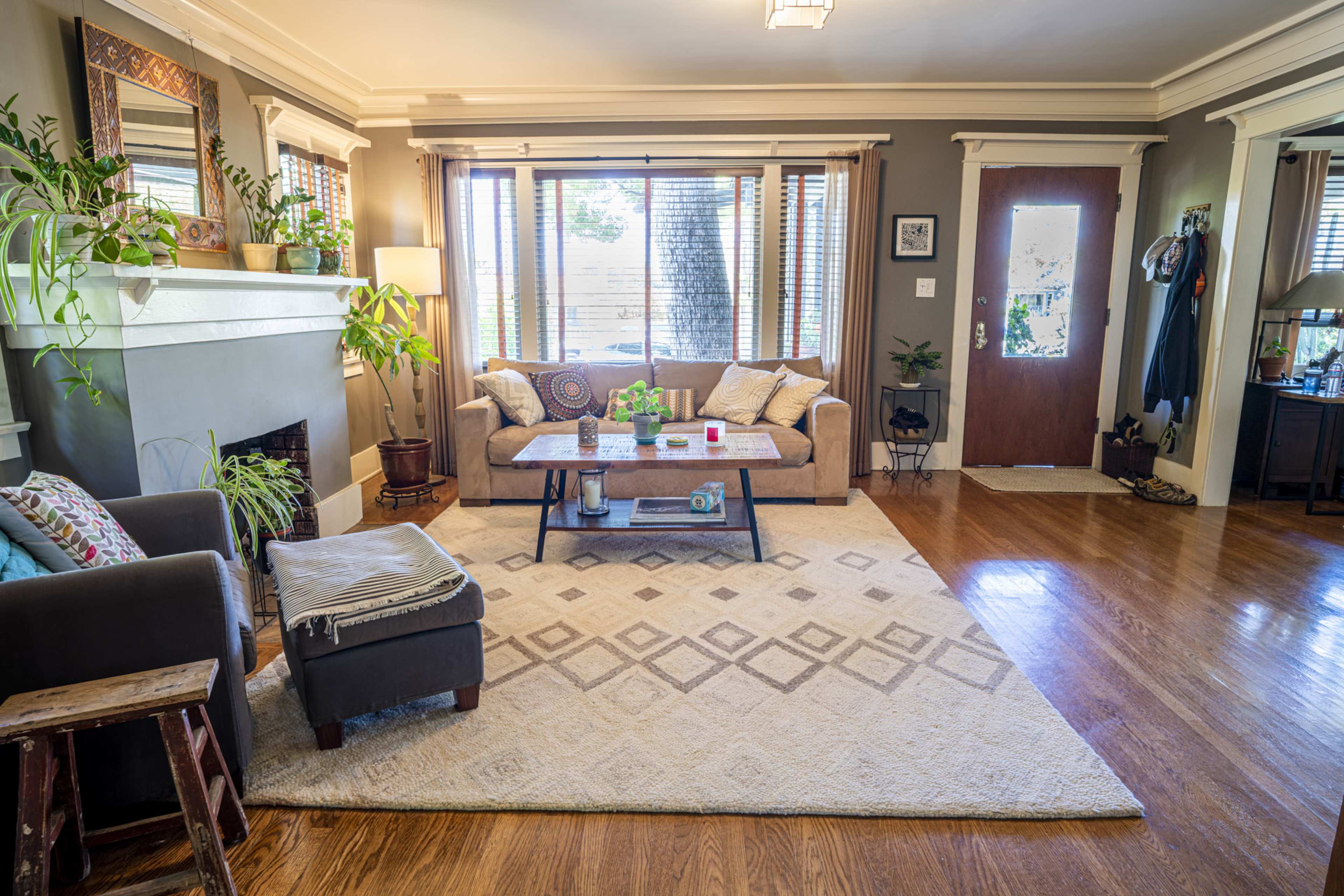 A living room features a sofa, coffee table, and large windows, with plants and a door leading outside.