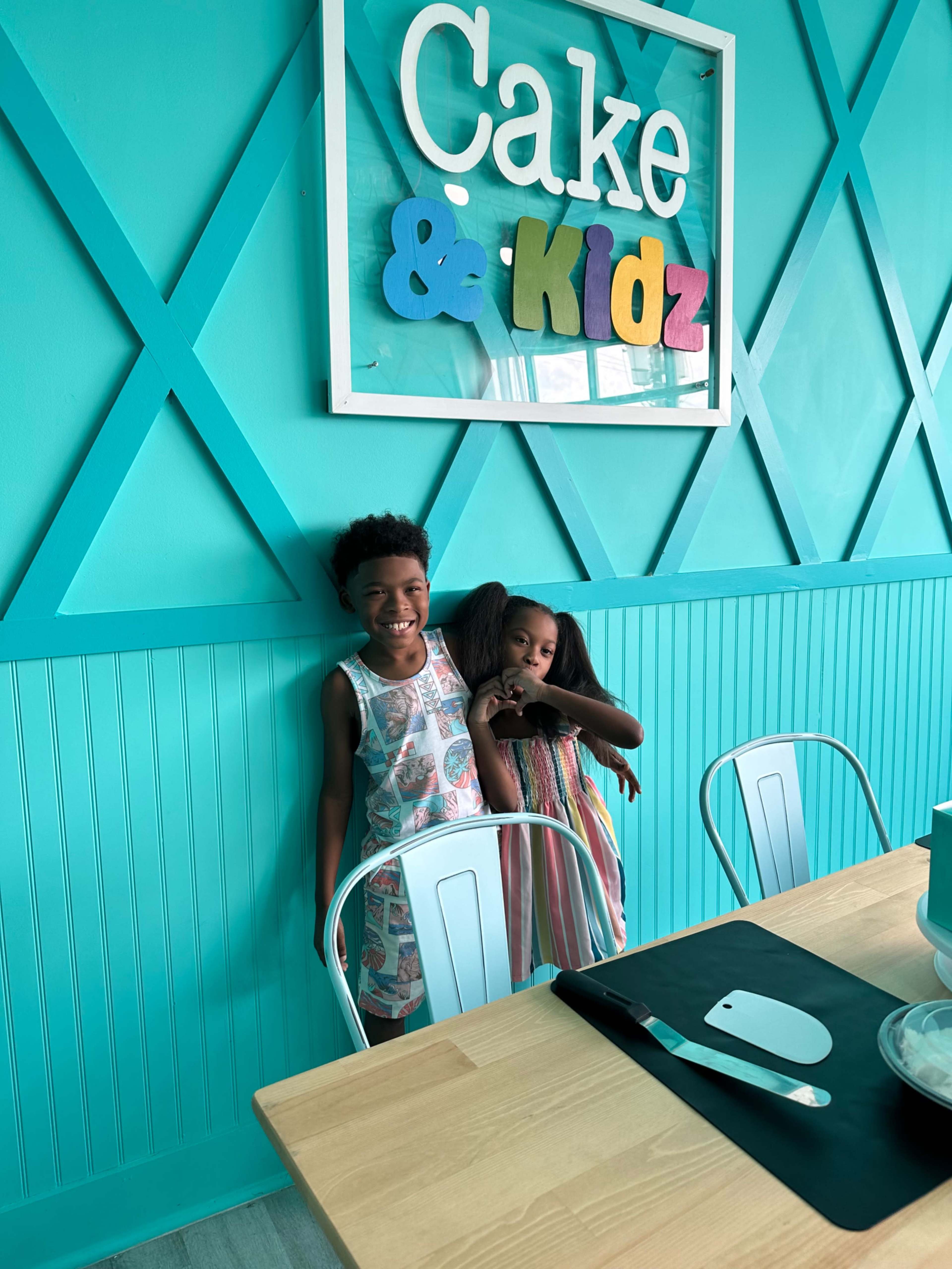 Two children stand beside a turquoise wall decorated with a sign that reads "Cake & Kidz."
