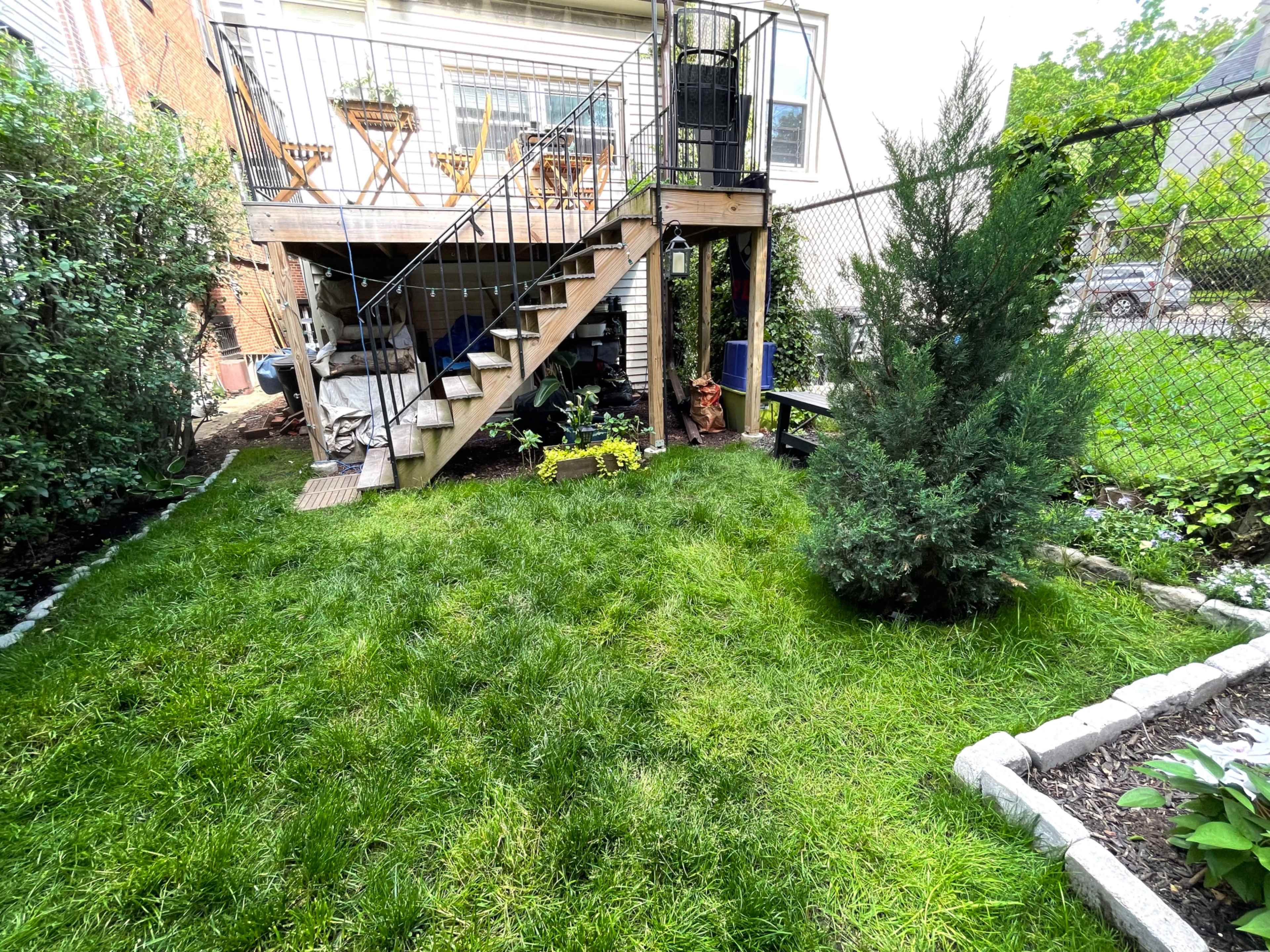 A grassy backyard with a staircase leading to a deck, plants, and a fence in the background.