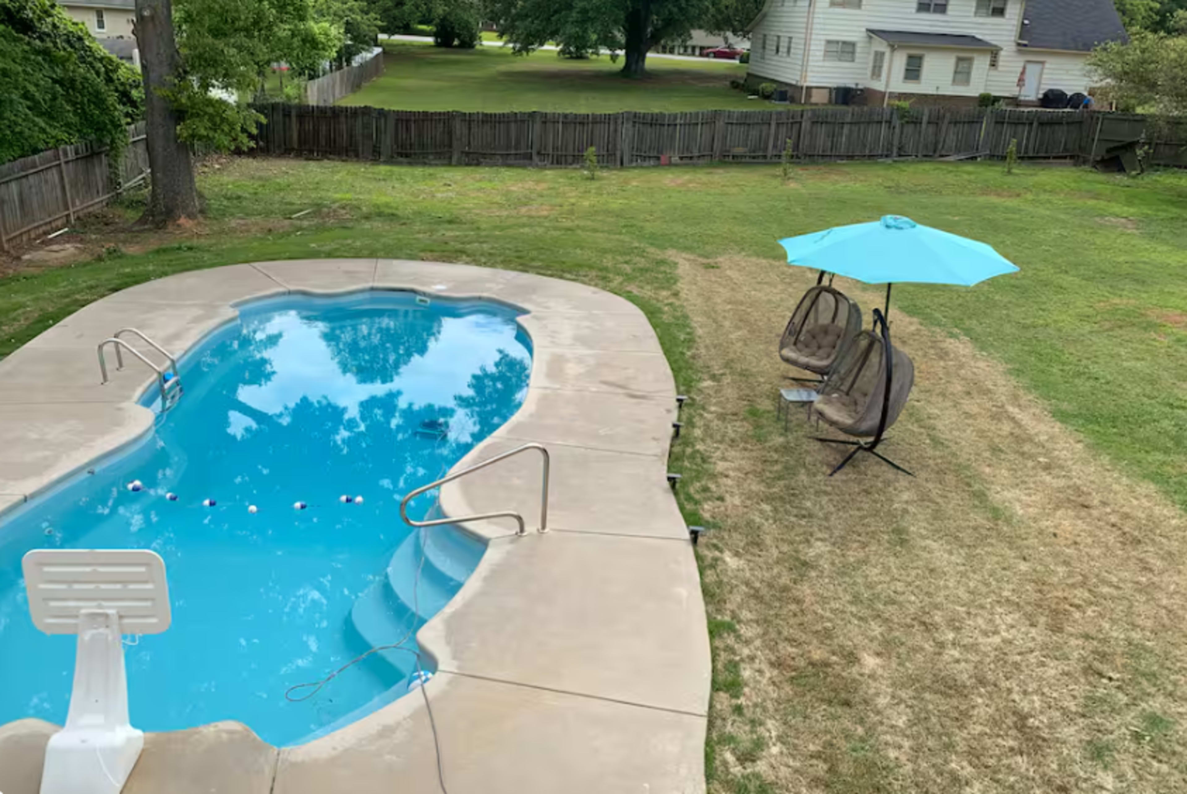 The image shows a backyard with a swimming pool, two hanging chairs under a light blue umbrella, and a grassy area with a wooden fence in the background.