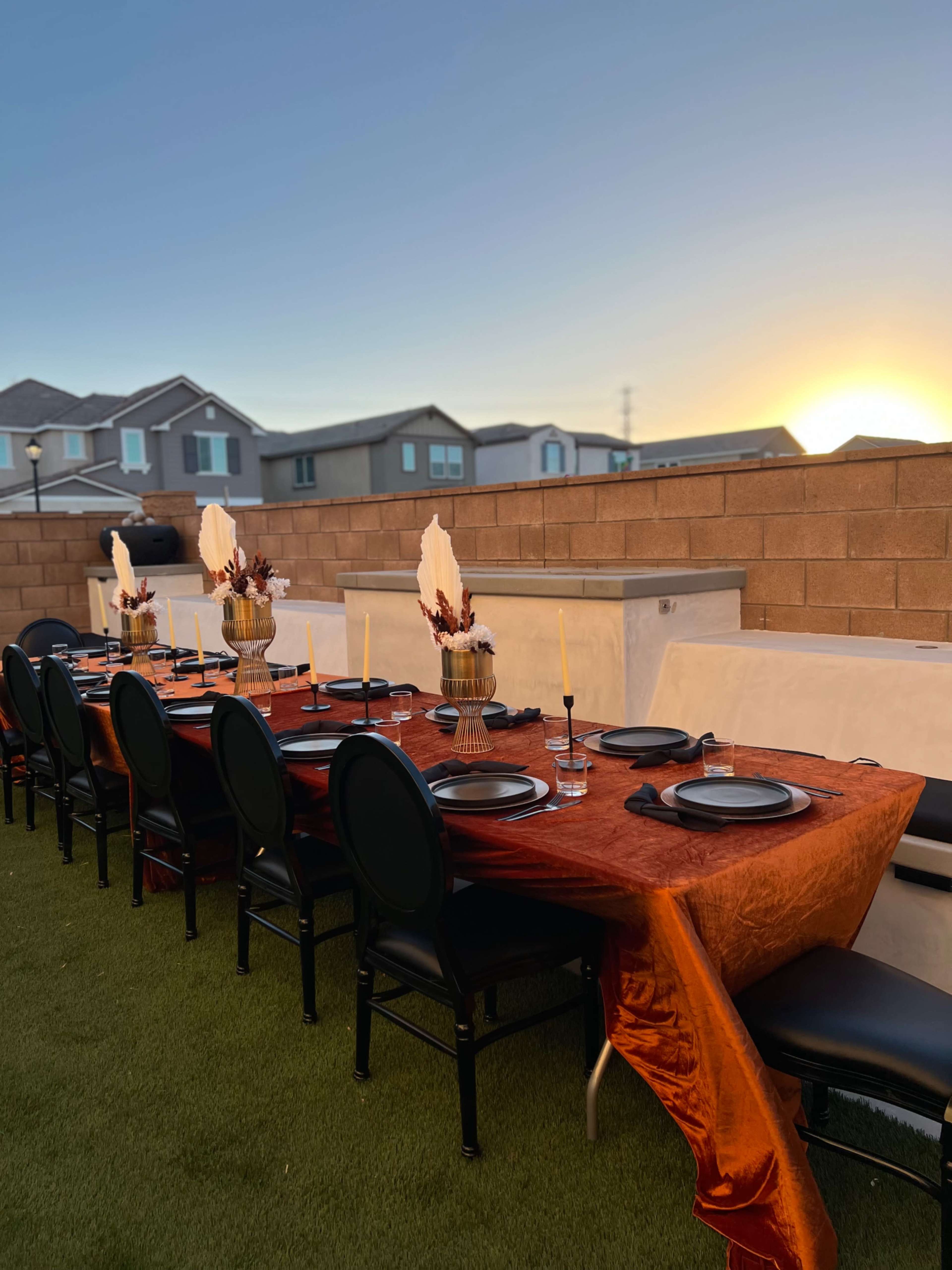 A long table is set with black chairs and an orange tablecloth, adorned with decorative centerpieces, against a backdrop of houses and a sunset.