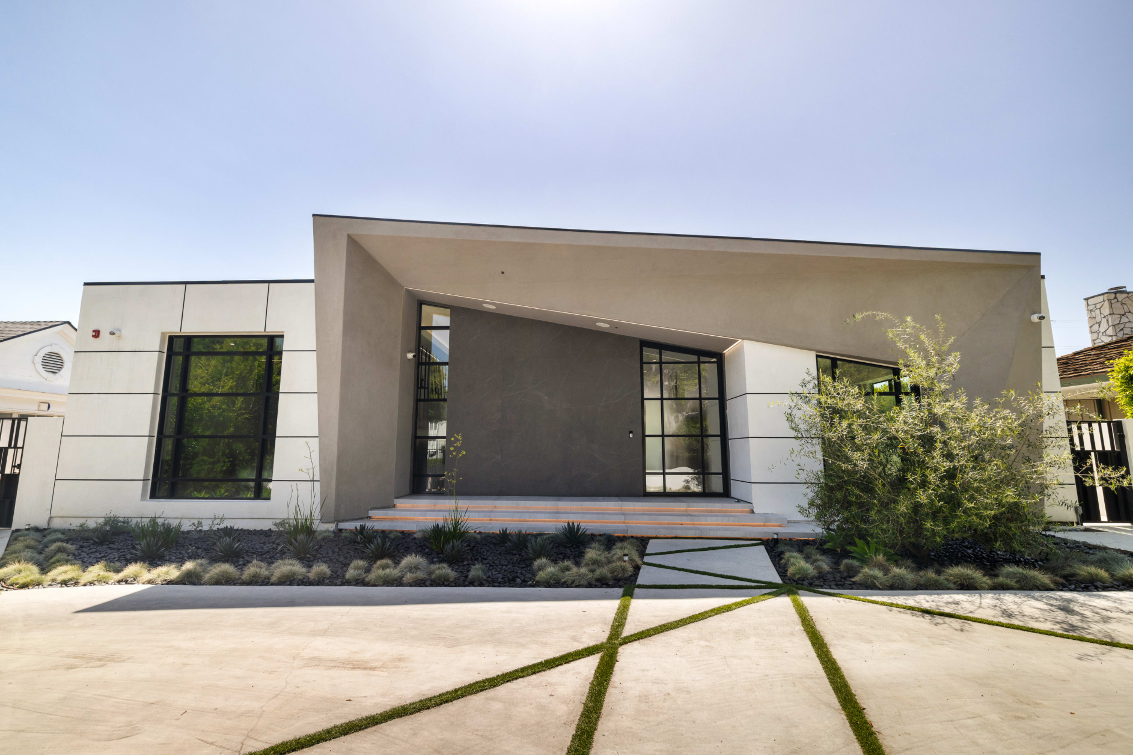 The image shows a modern house with an angular design, featuring large windows and a landscaped front yard.